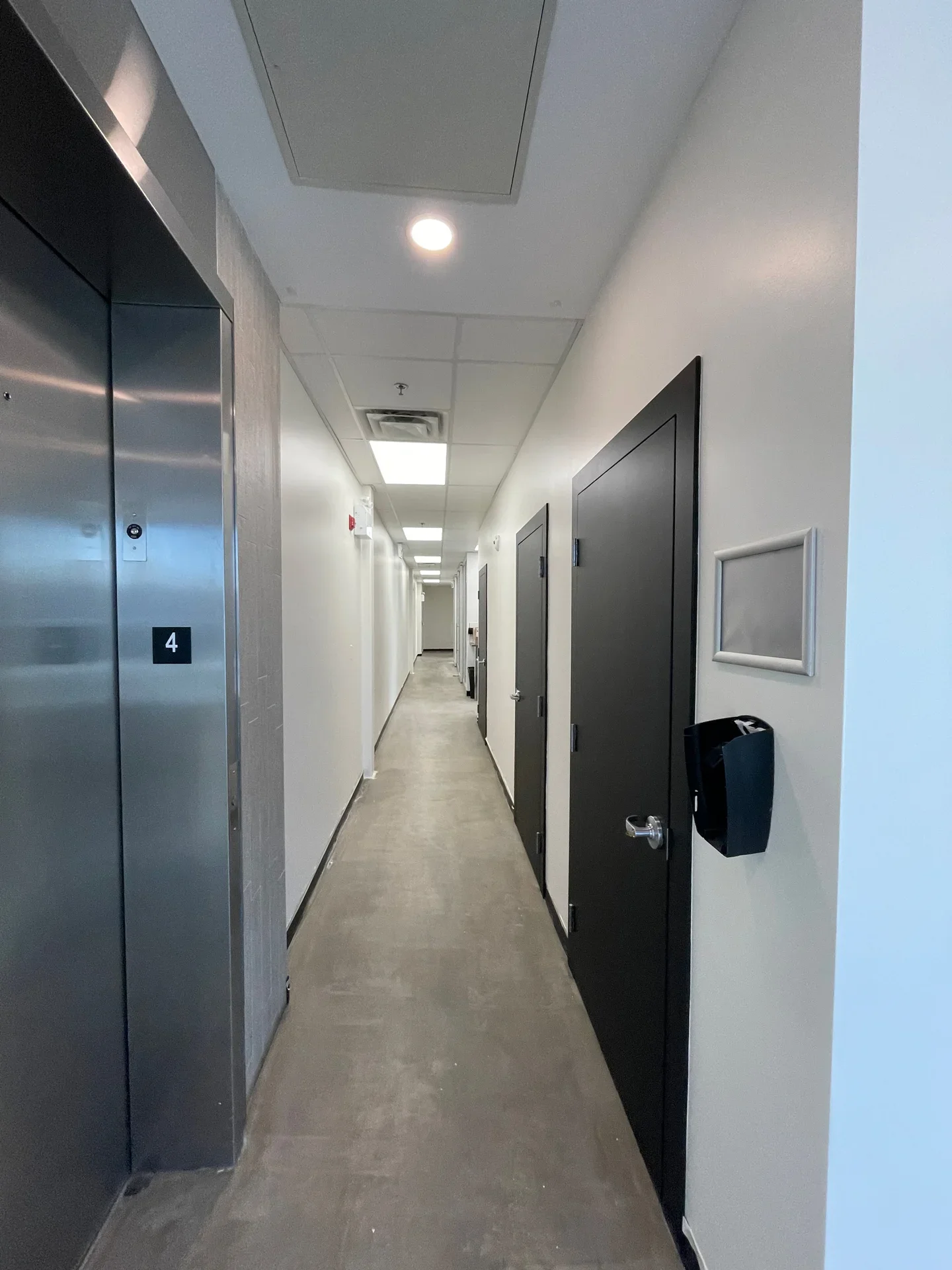 Long, narrow hallway in an office building with gray and white walls, black doors on the right, an elevator with stainless steel doors on the left, and ceiling lights.