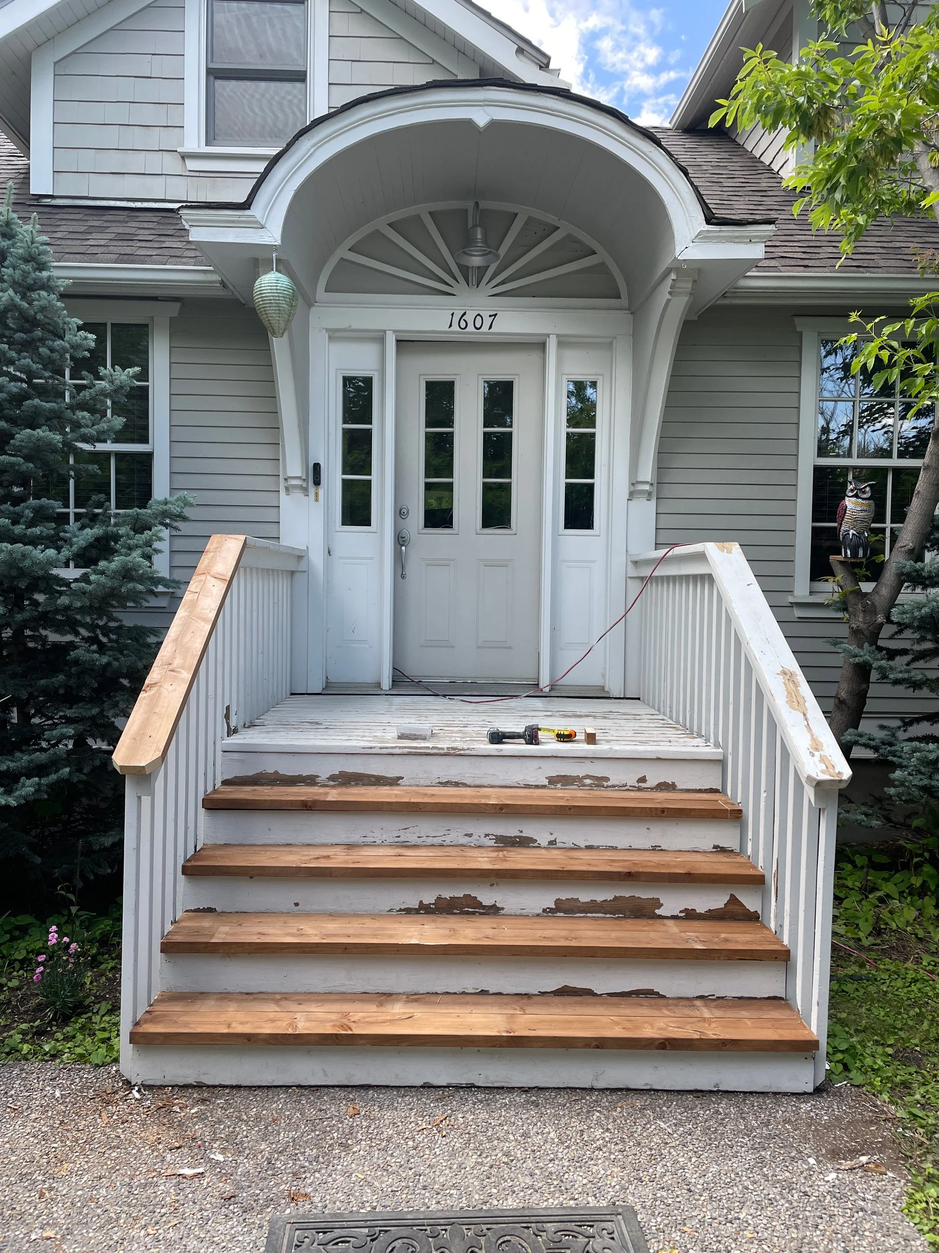 Front porch steps of a house under renovation, with some steps newly replaced with wood and others still old and worn. Tools are on the porch, and the house number 1607 is visible above the white front door.