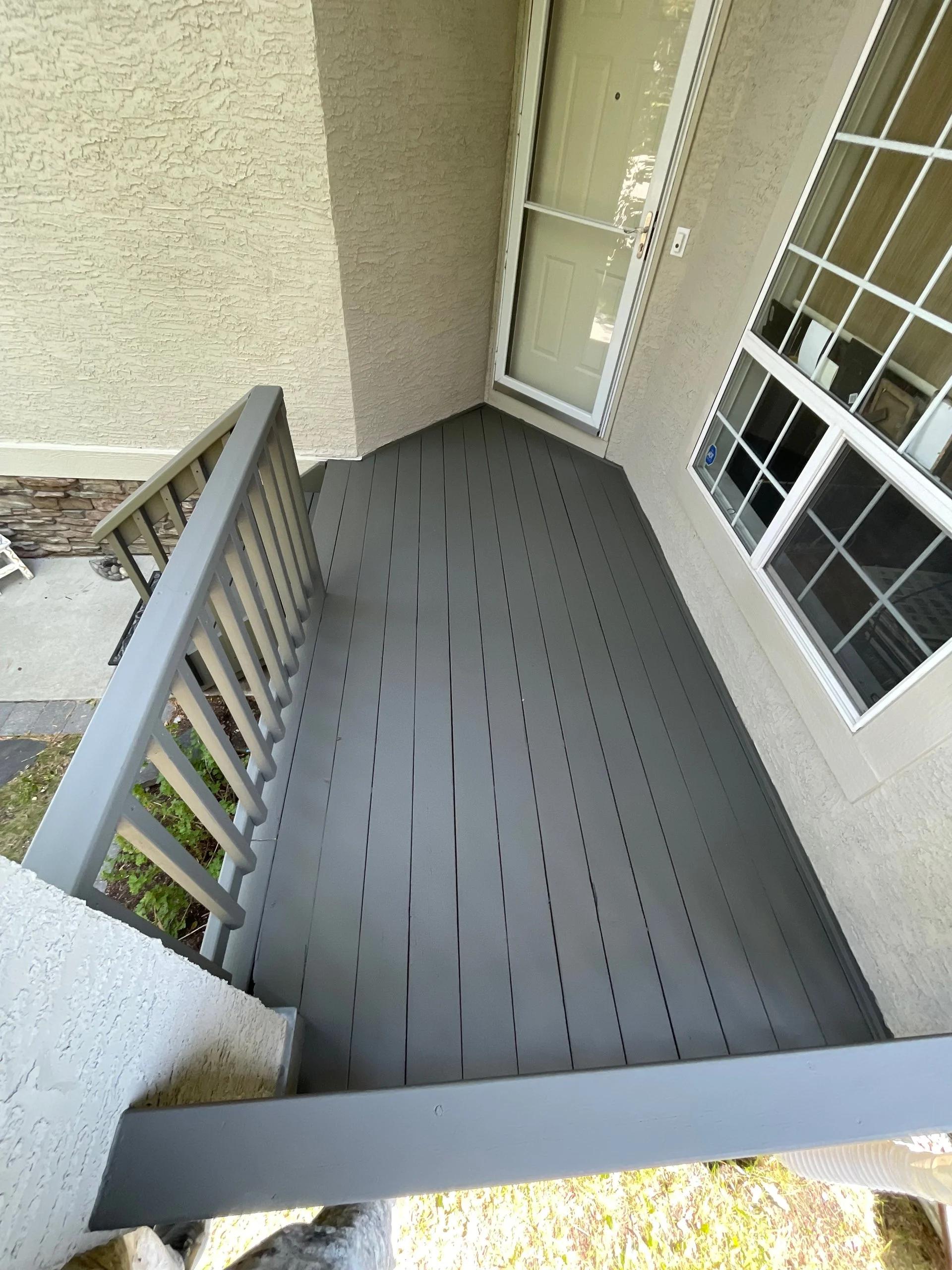 Small backyard balcony with gray wood flooring, gray railing, beige exterior walls, glass door, and large window.