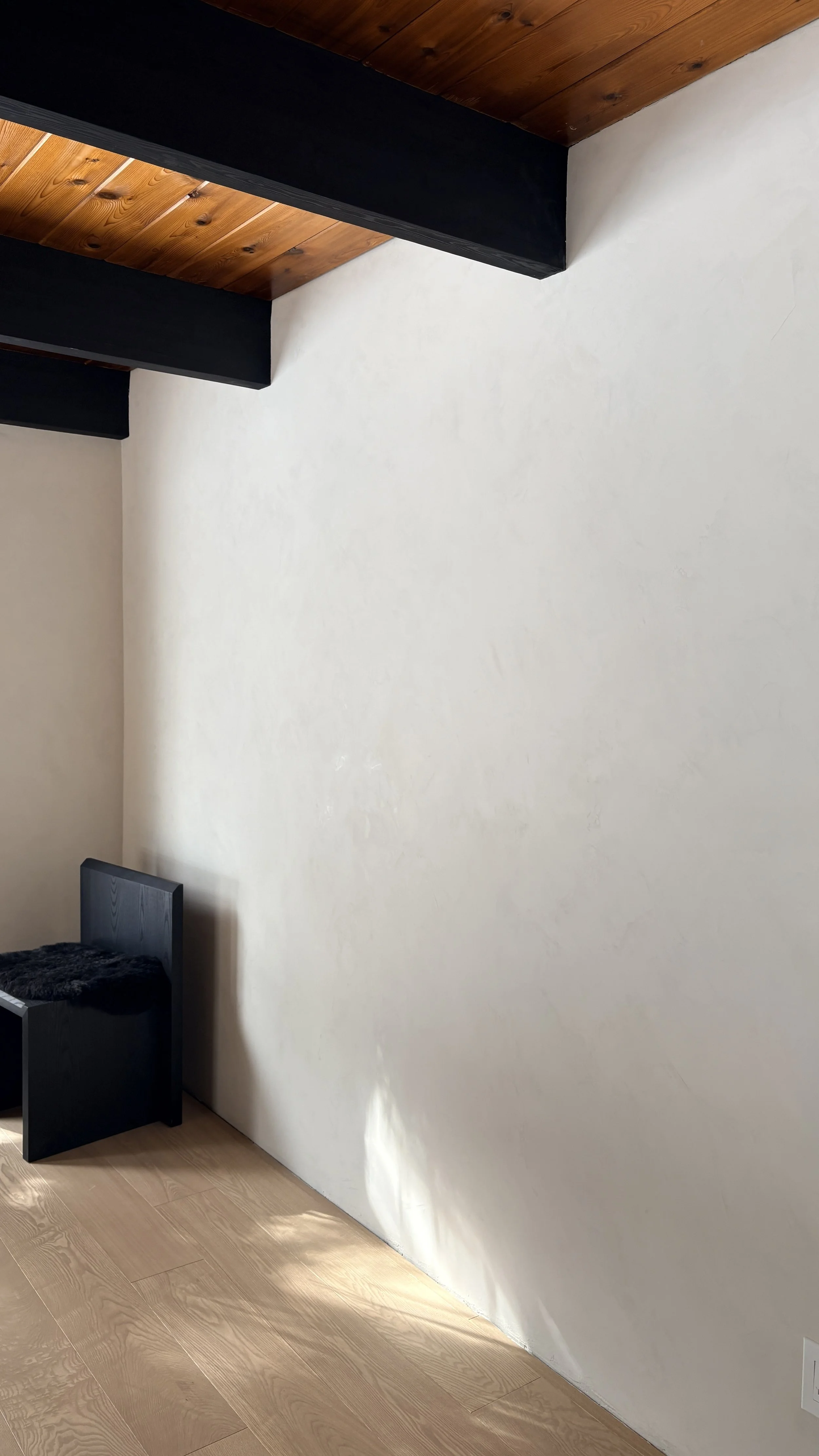 A corner of a room featuring a white textured wall, wooden ceiling with exposed beams painted black, a light wooden floor, and part of a black bench with a fur cushion.