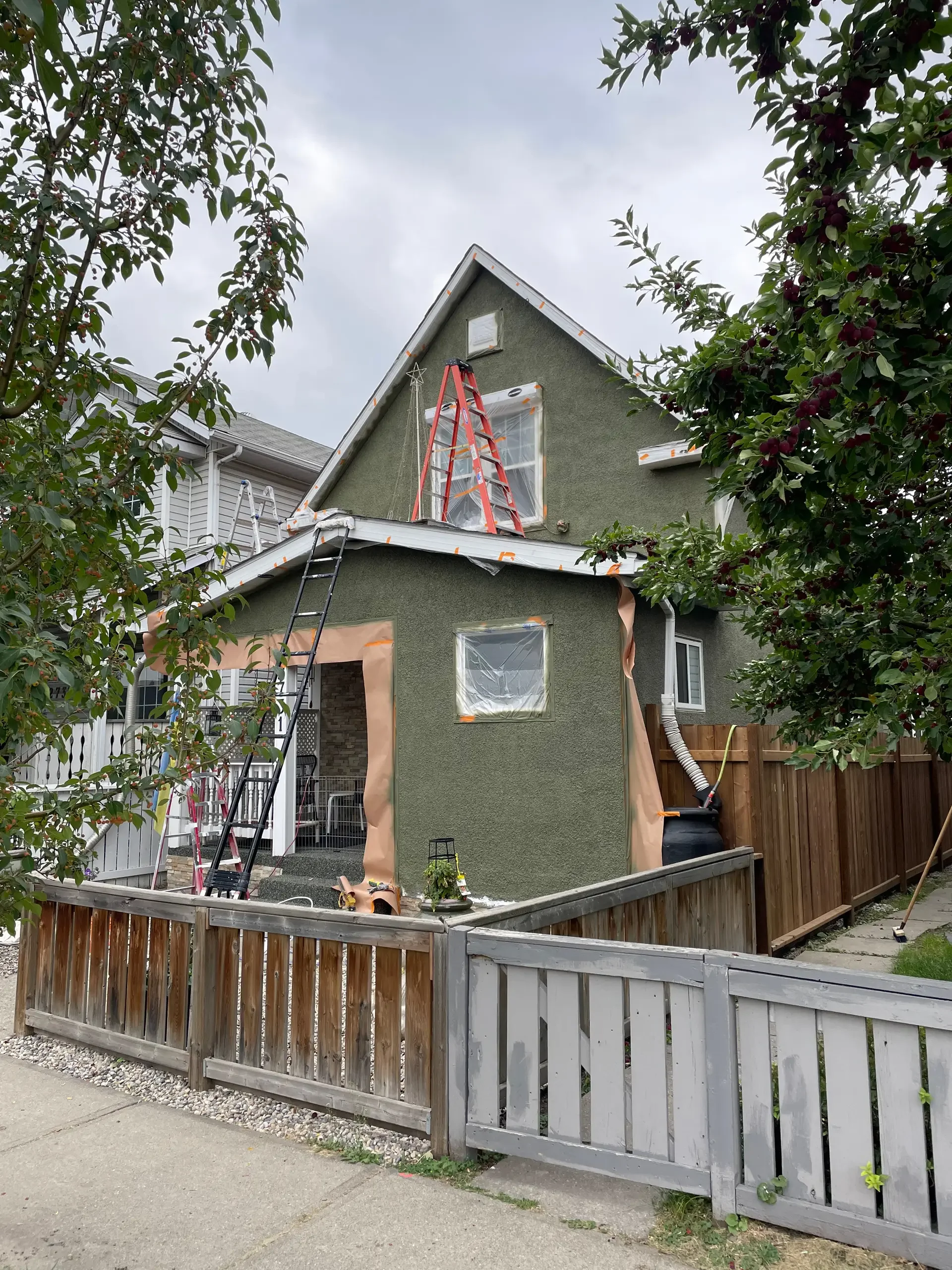 House under renovation with plastic sheeting over windows, ladders leaning against the house, and tape around the roof edges, surrounded by trees and a wooden fence.