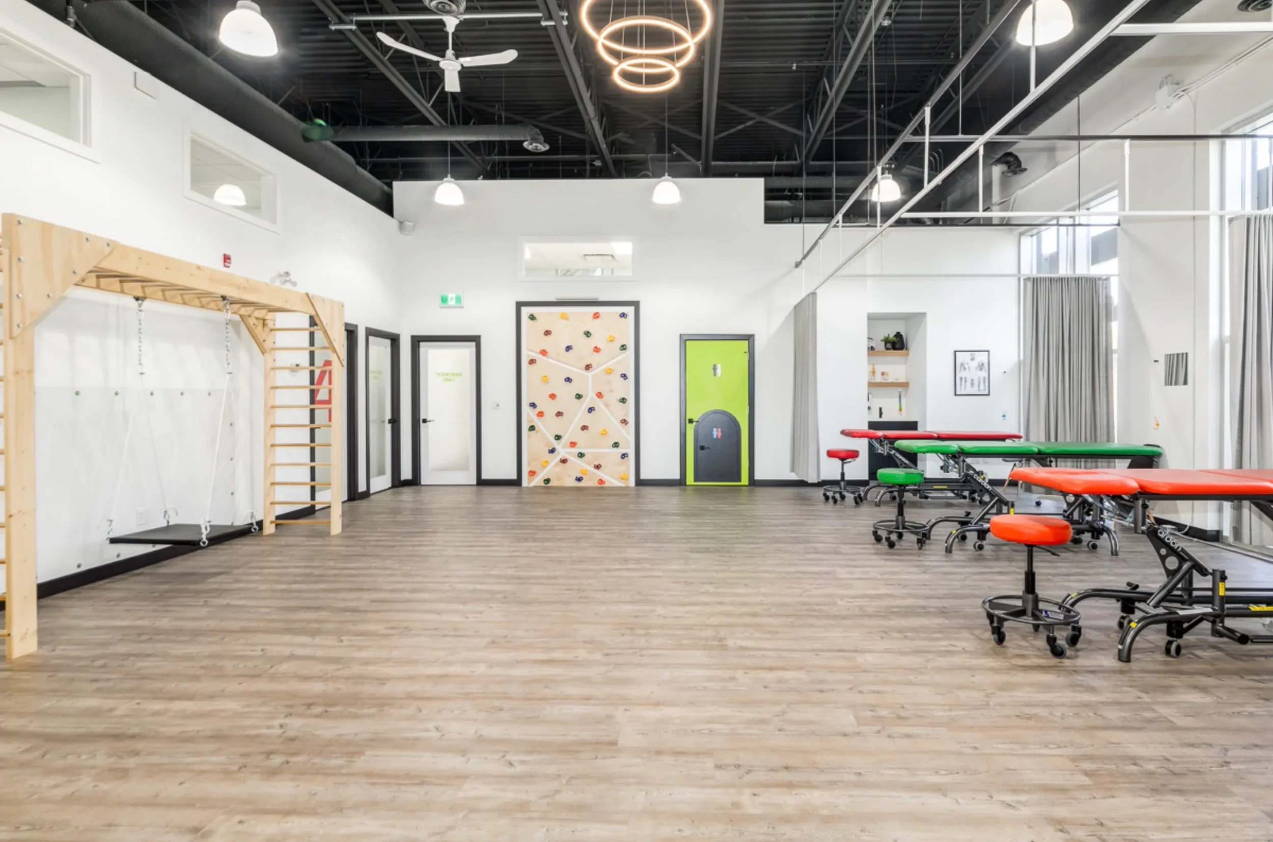 Empty physical therapy or rehabilitation room with wooden floor, colorful tables, chairs, and equipment for exercises and therapy.