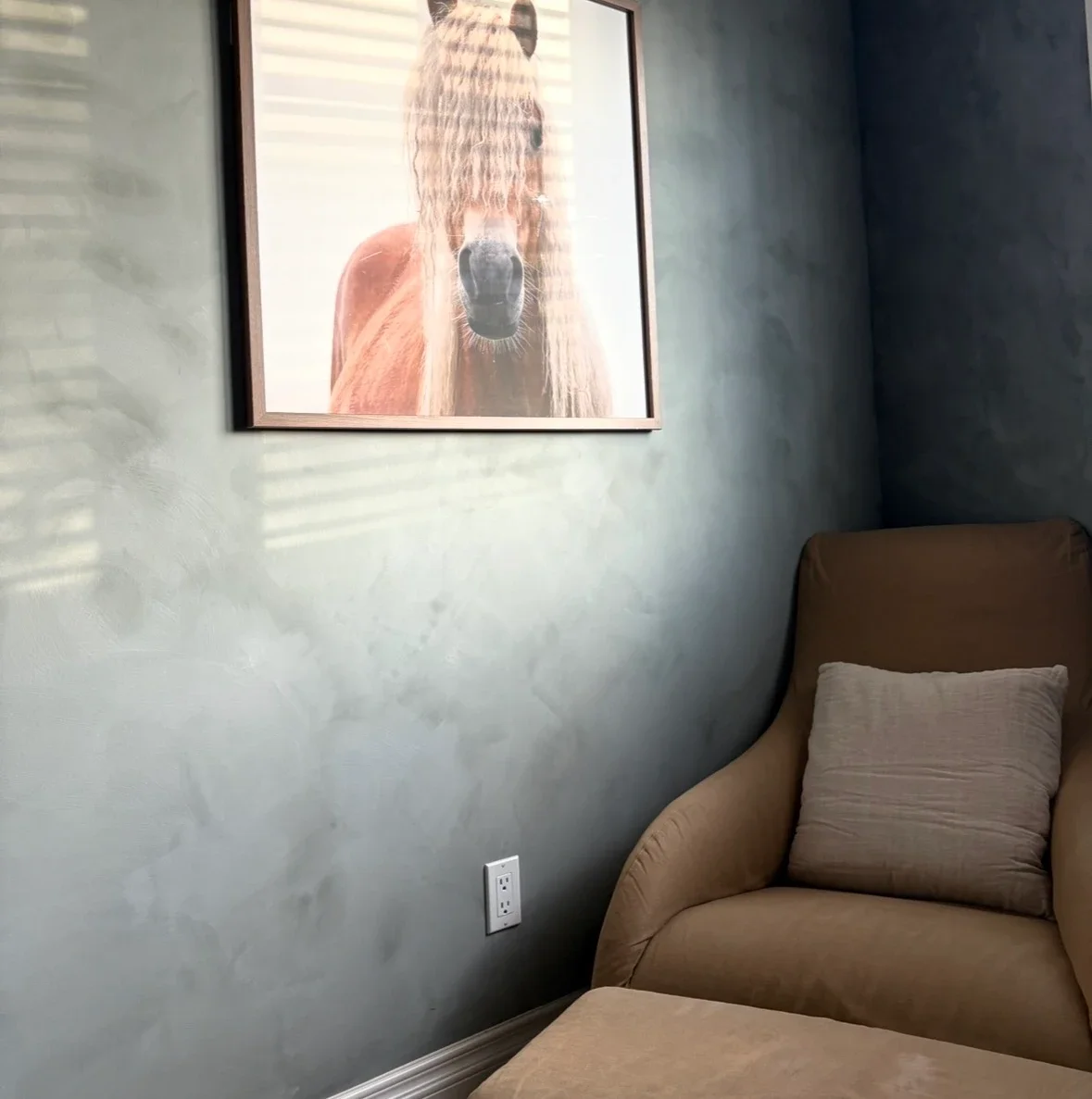 A living room corner features a framed horse portrait hanging on a wall, with sunlight filtering through blinds casting shadows. A beige armchair with a matching pillow is positioned nearby.