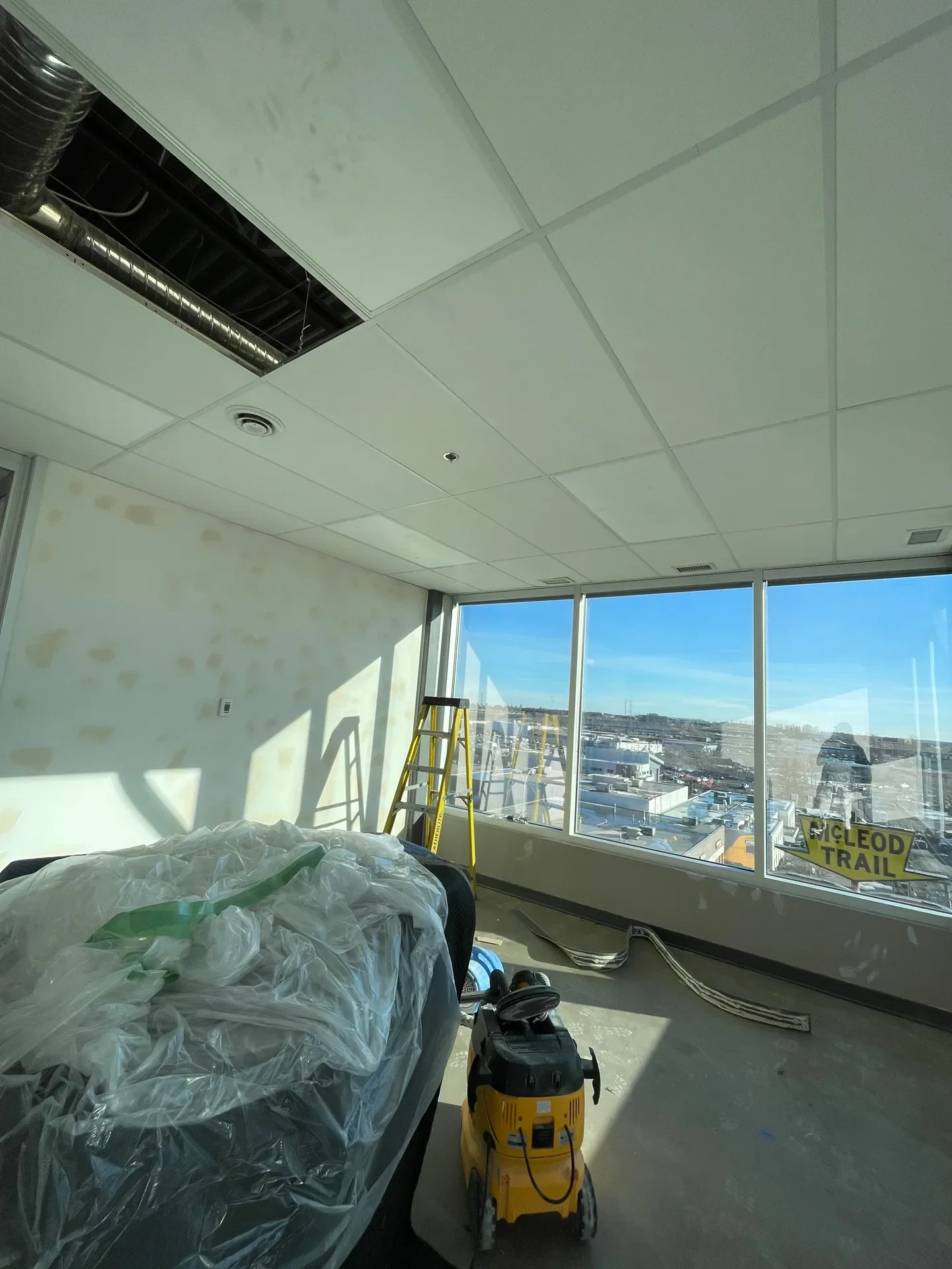 Construction site inside an office building with large windows, a yellow ladder, construction debris, and a yellow air compressor.