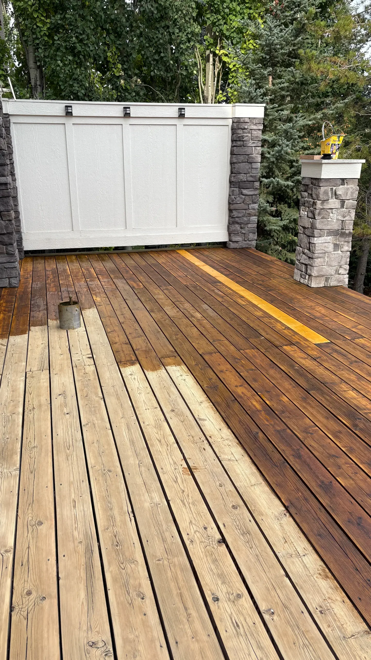 A wooden deck with partially stained planks, white fencing with black hardware, and stone pillars, surrounded by trees.