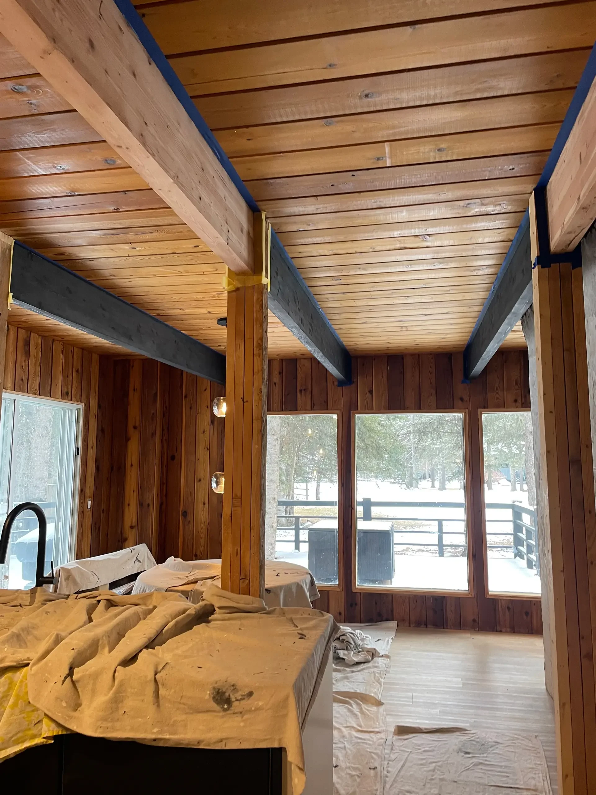 Interior view of a wooden cabin under construction, with visible ceiling beams and large windows showing a snowy outdoor landscape.