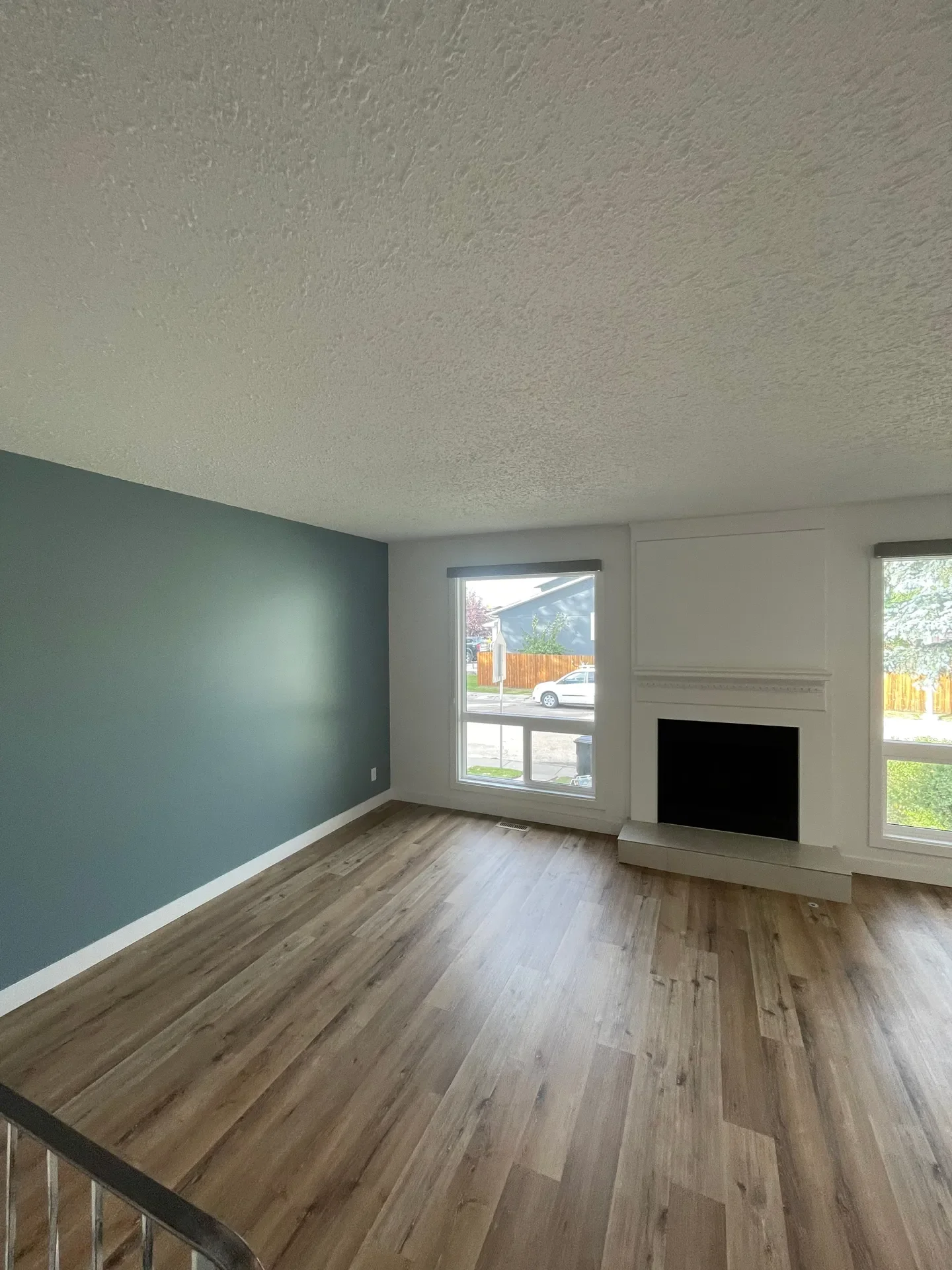 Empty living room with a teal accent wall, hardwood floors, a fireplace, and two large windows.