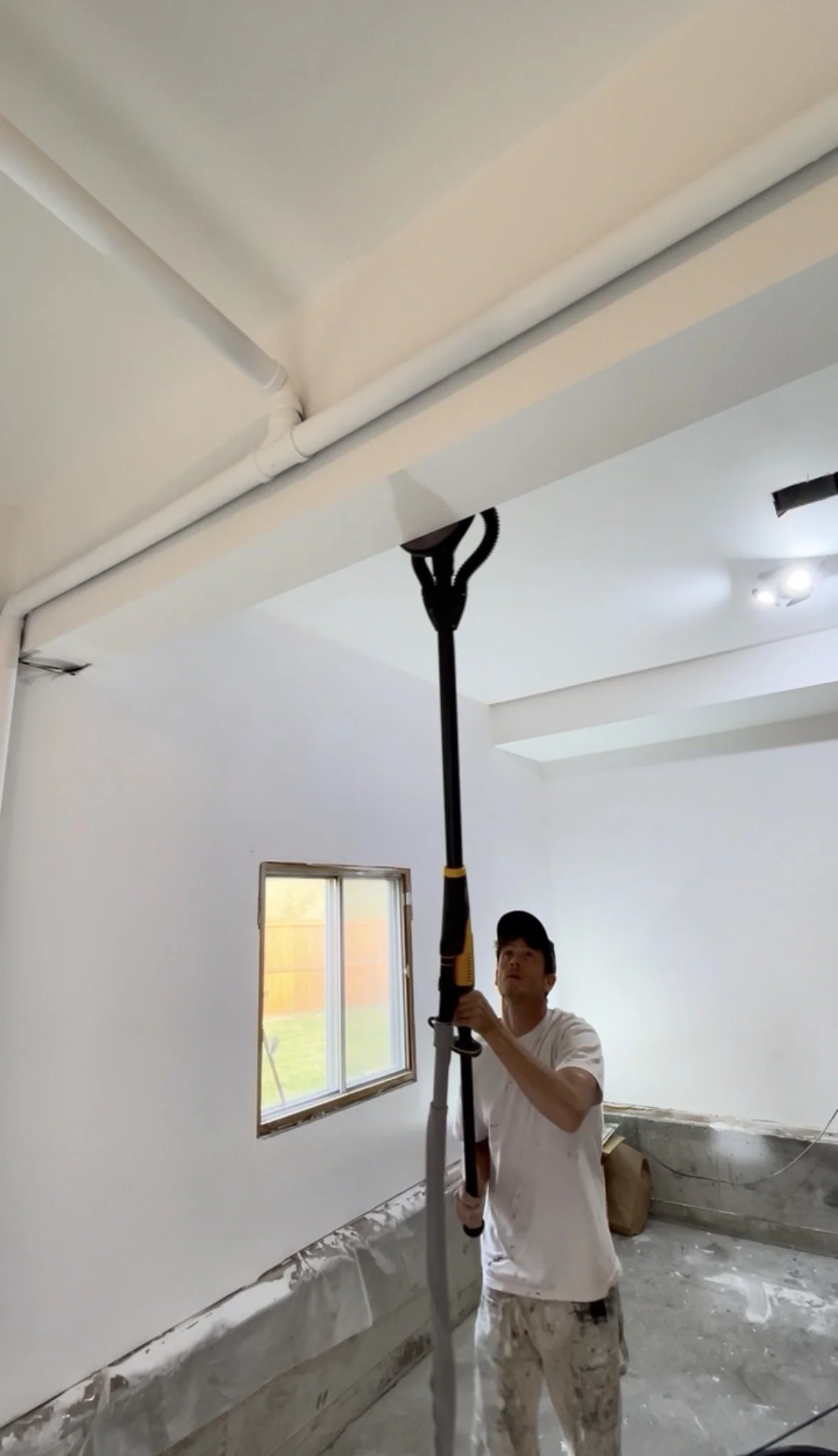 A man in white paint-splattered clothes holding a long tool reaching the ceiling in an unfinished room with white walls, a window, and construction materials on the floor.