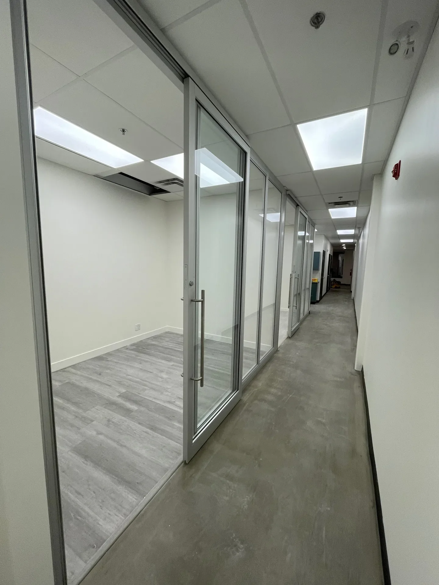 Empty office room with glass walls and door, next to a hallway with industrial flooring and ceiling lights.