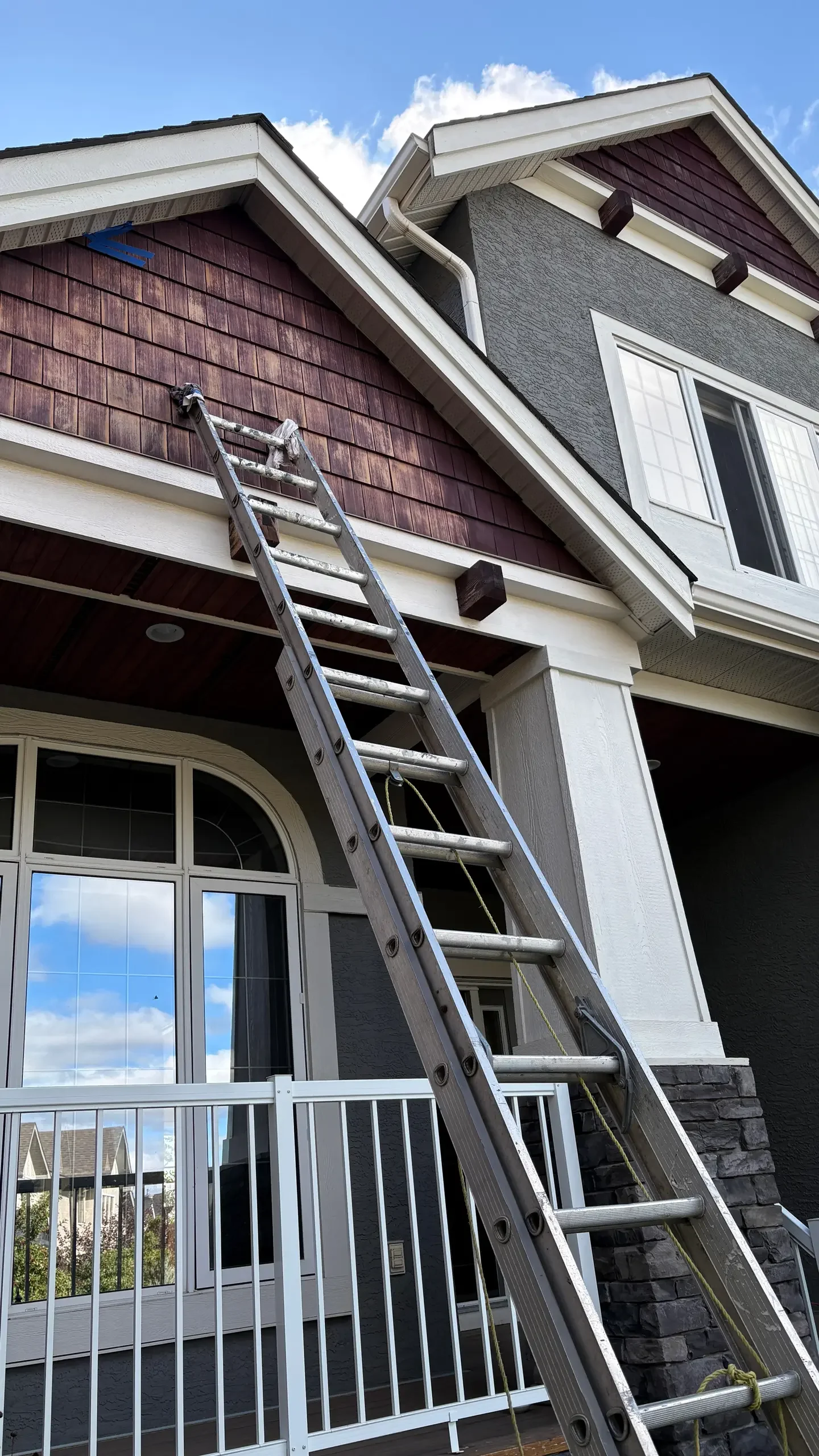 A multi-story house with gray and brown exterior walls, a ladder leaning against the front, and a porch with a white railing.