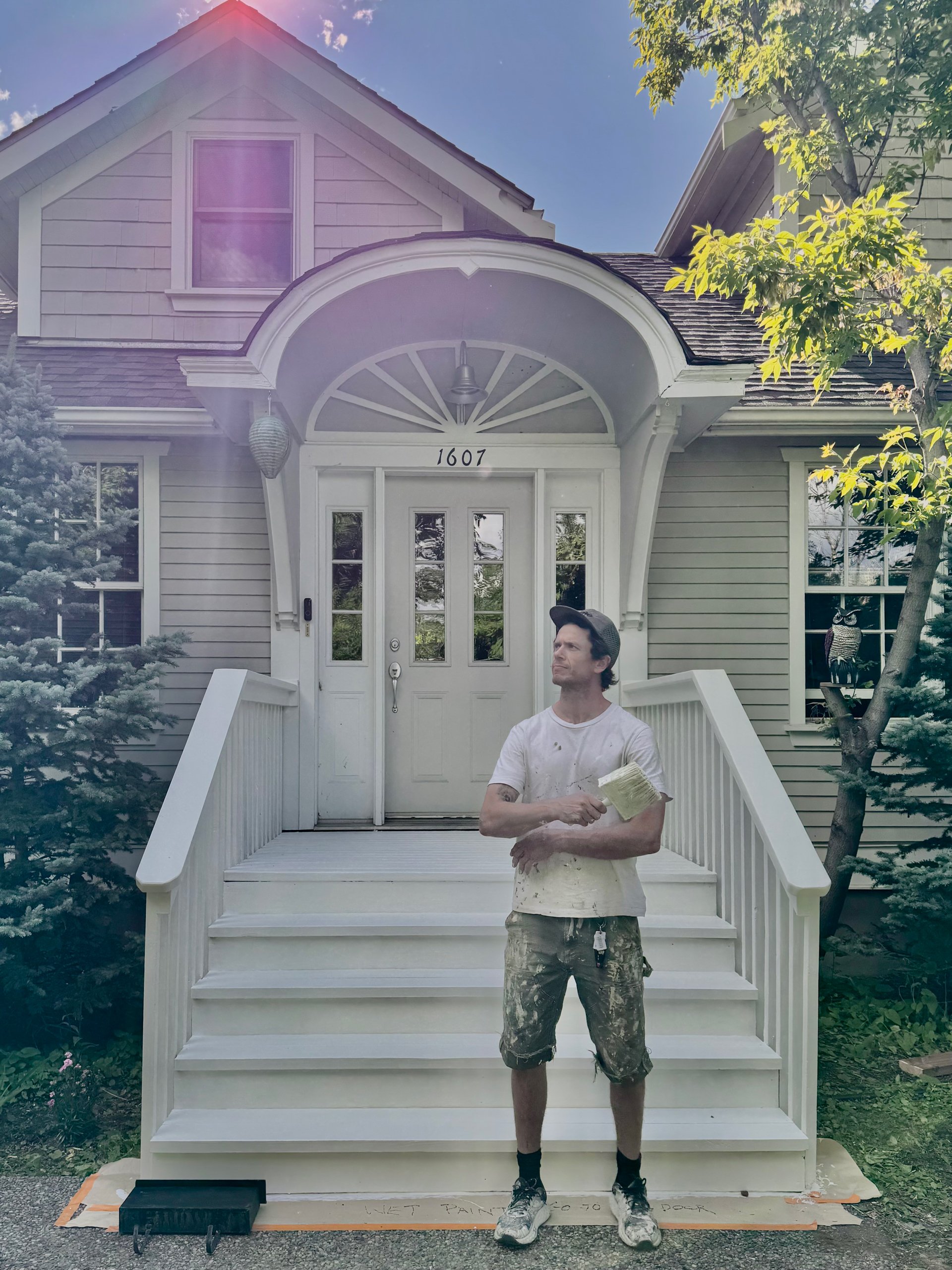 A man standing on the porch of a house, holding a paintbrush and wearing paint-splattered clothes. The house has a white front door, steps, and surrounding greenery.