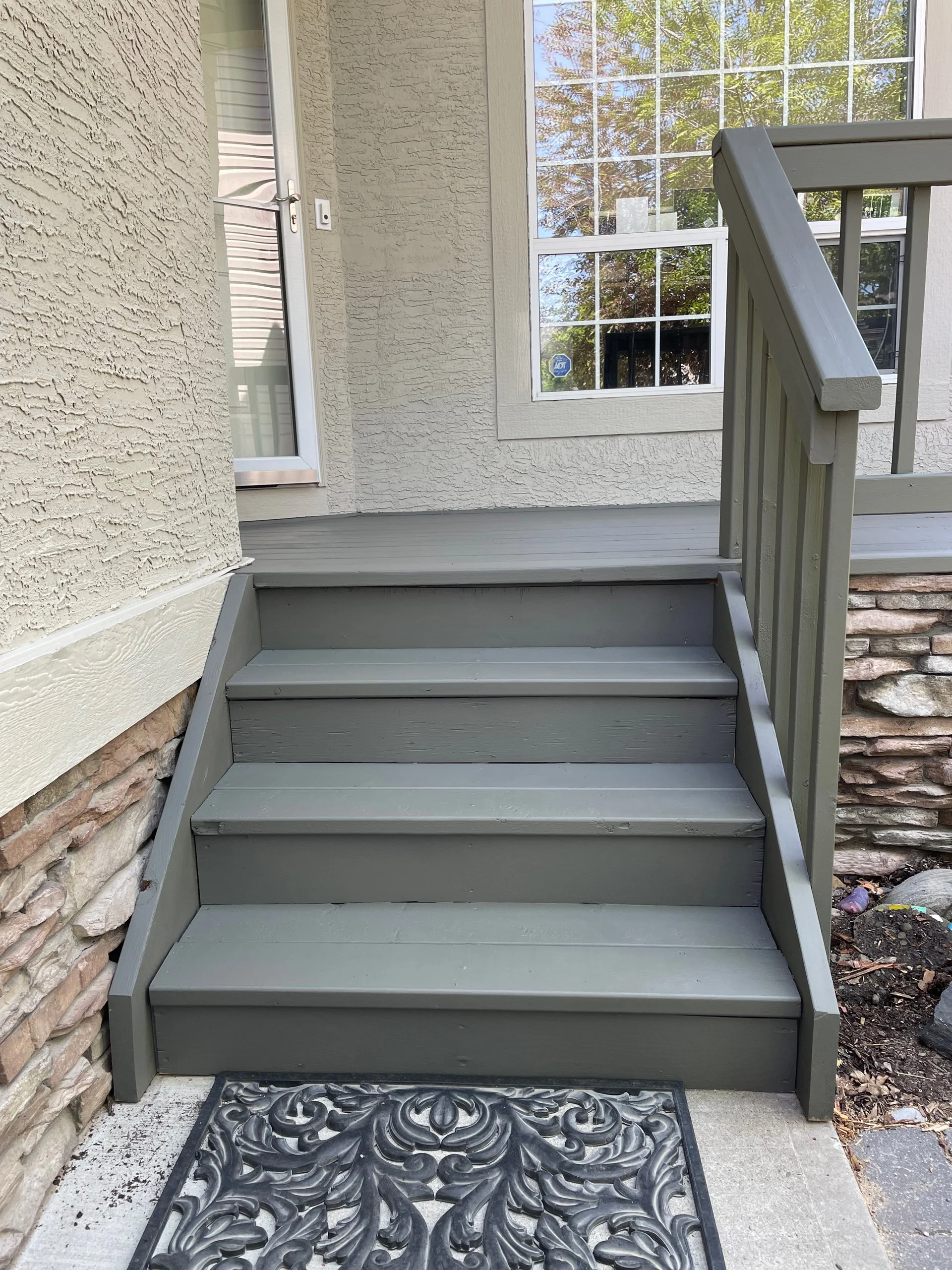 A small set of gray wooden stairs leading up to a porch with a large window and a white door with a glass panel. There is a decorative black and gray doormat at the bottom of the stairs.