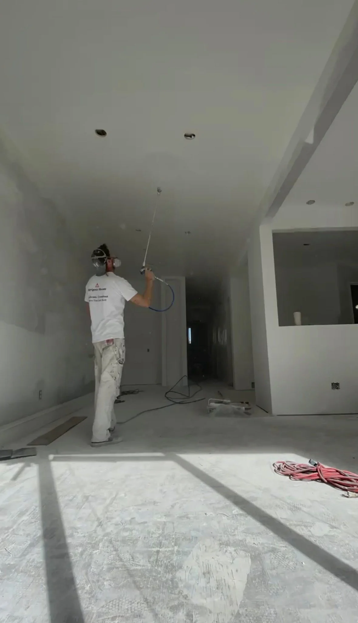 A construction worker wearing a white shirt, jeans, and a helmet is using a spray gun to paint the ceiling of a room under construction.