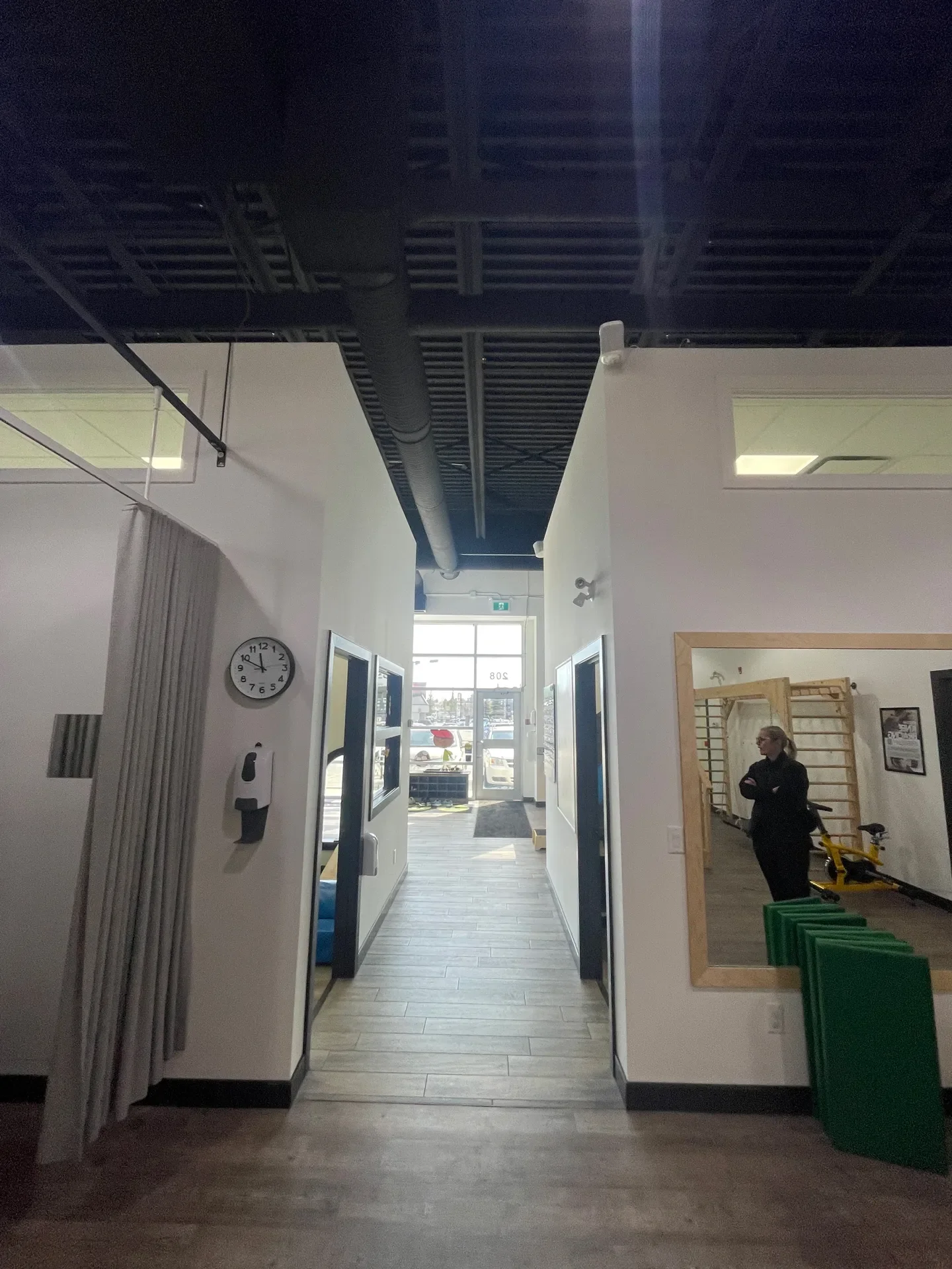 Interior of a modern gym or fitness center with a corridor leading to the entrance with glass door, a person standing in front of a mirror, and exercise equipment visible in the background.