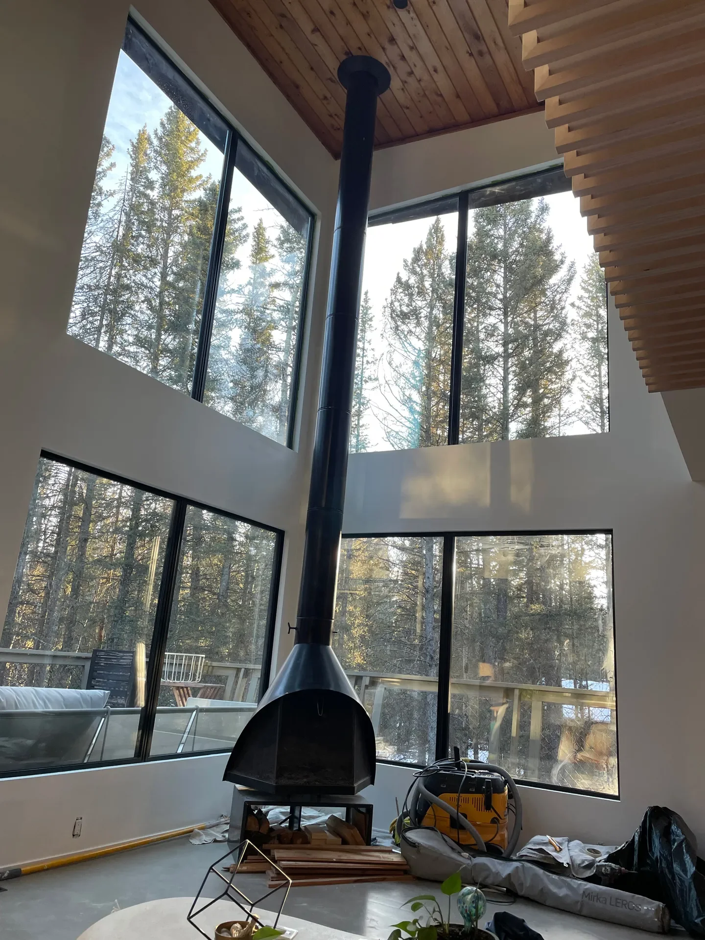 Interior of a modern house with large floor-to-ceiling windows showing a forest view. There is a black fireplace pipe extending from the floor to the ceiling, with construction tools and materials scattered on the floor.