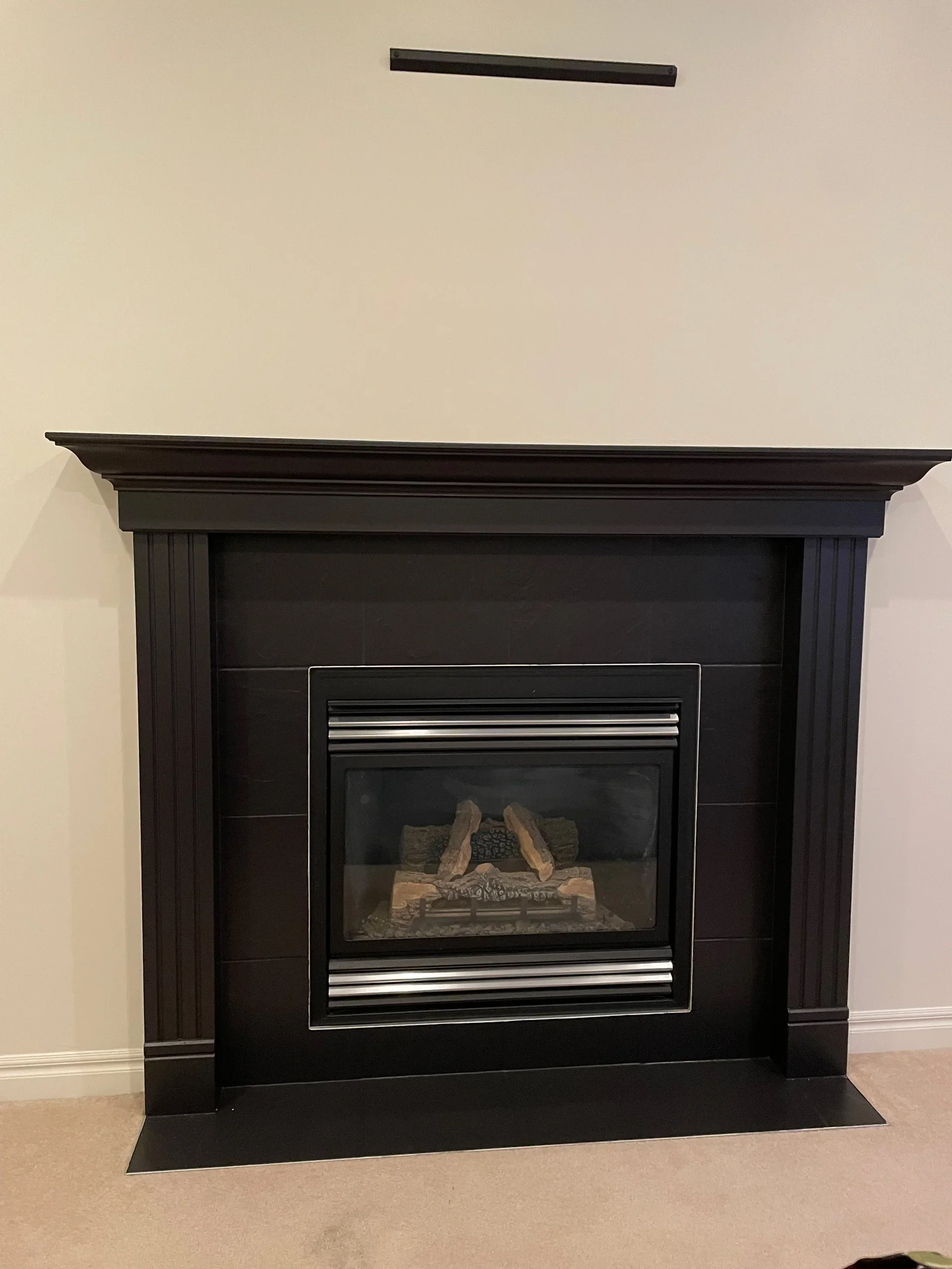 Black fireplace with a mantel and a glass cover, inside are decorative logs and stones, with a beige carpet beneath and a plain light-colored wall.