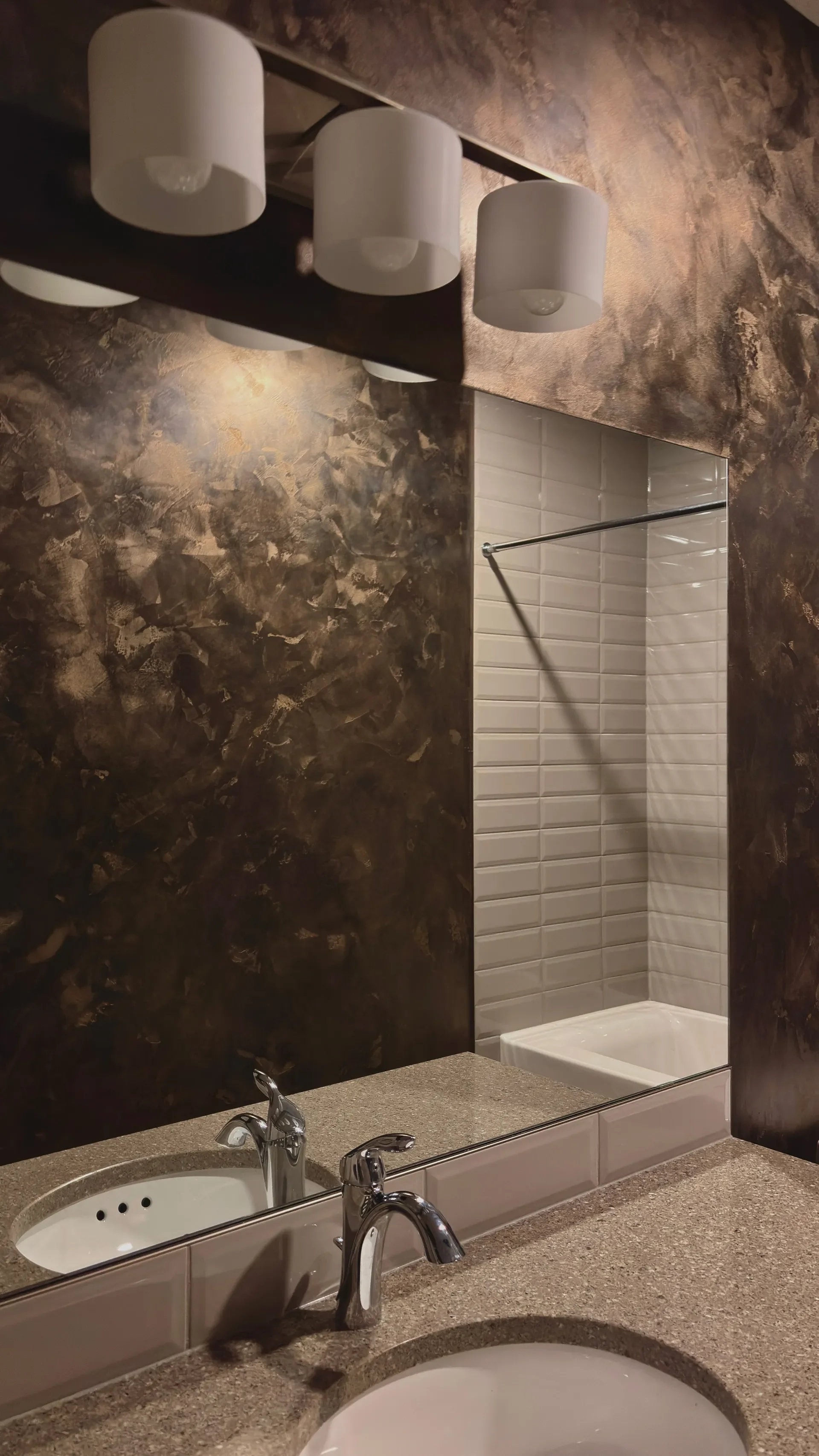 Bathroom mirror above a sink with stone countertop, two faucets, and a large mirror reflecting a shower area with white tiles and a curtain rod. Overhead, there are three white cylindrical light fixtures.