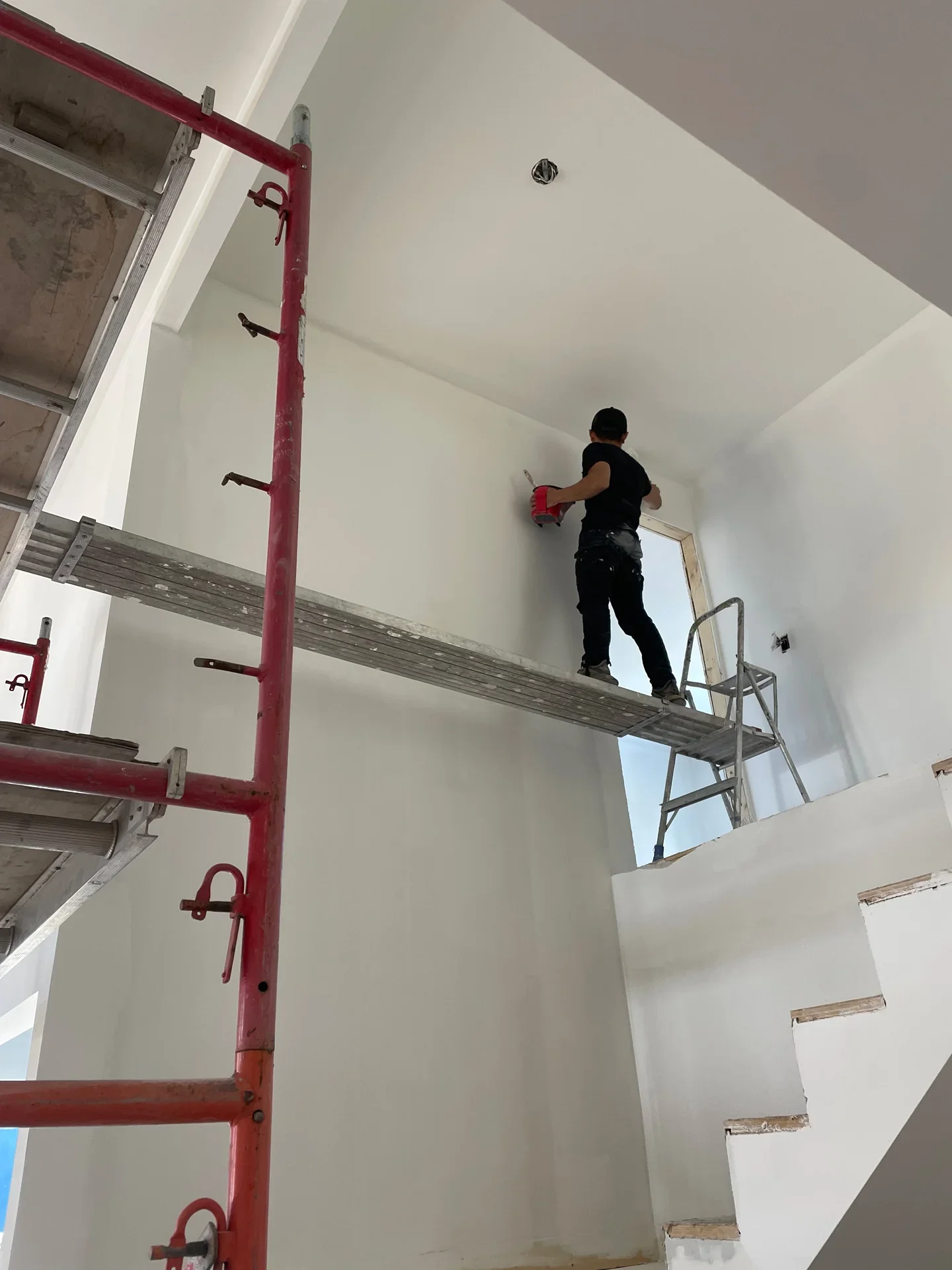 A person standing on scaffolding inside a house, painting the upper corner of a wall. There is a ladder leaning against the wall and an unfinished staircase.