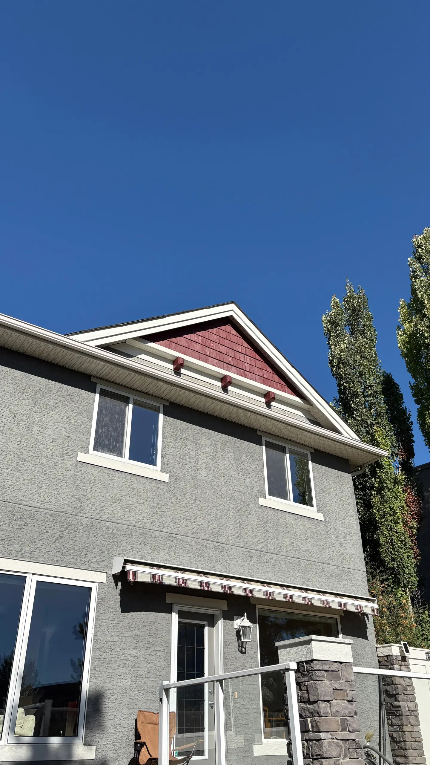 Front view of a gray two-story house with white trim, windows, a door with a striped awning, and a glass railing on the patio. The sky is clear and blue, with trees visible on the right side.