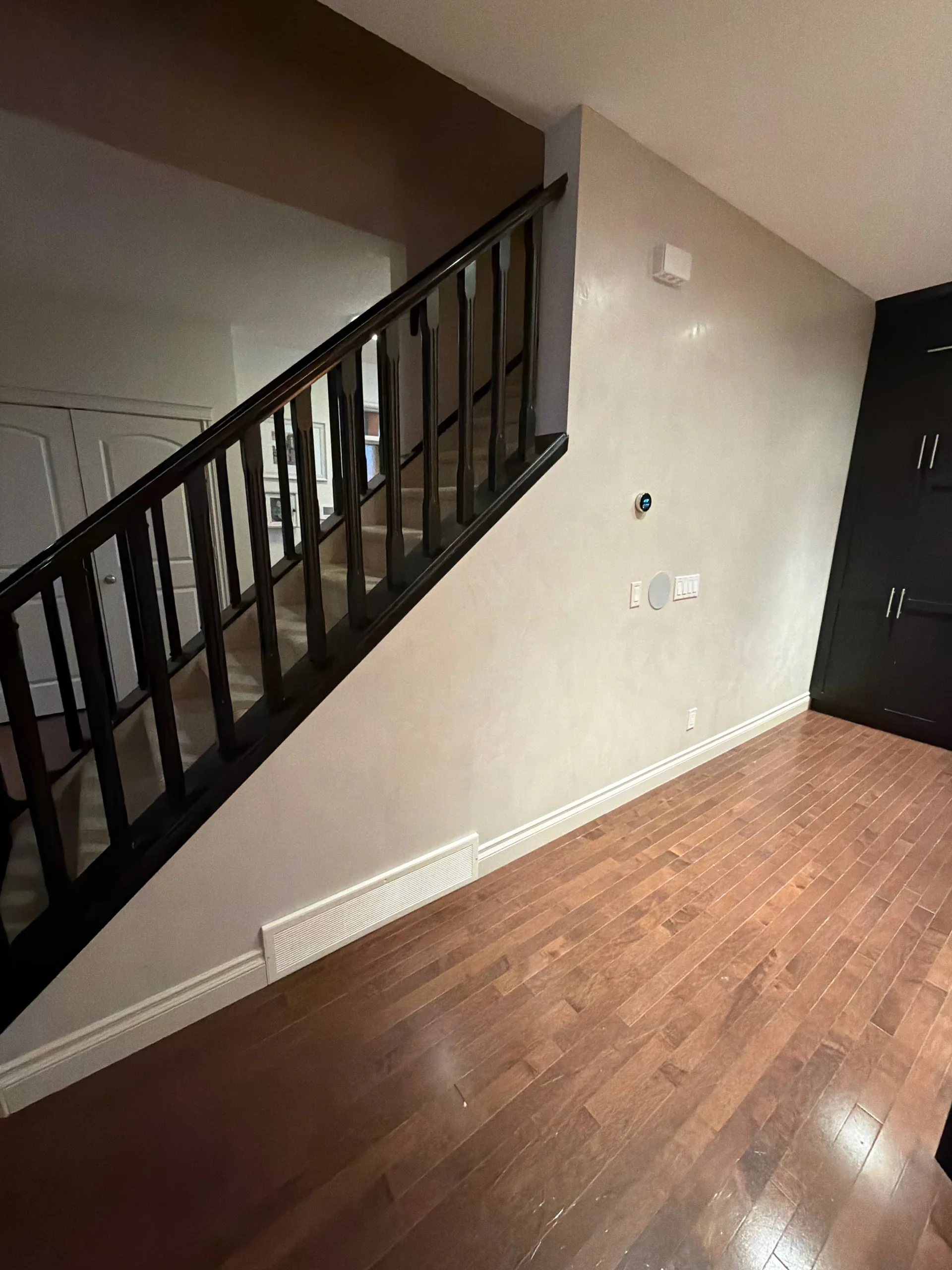 Interior view of a room with a staircase, hardwood floor, white walls, and a black cabinet. There are various electrical outlets and a thermostat on the wall.