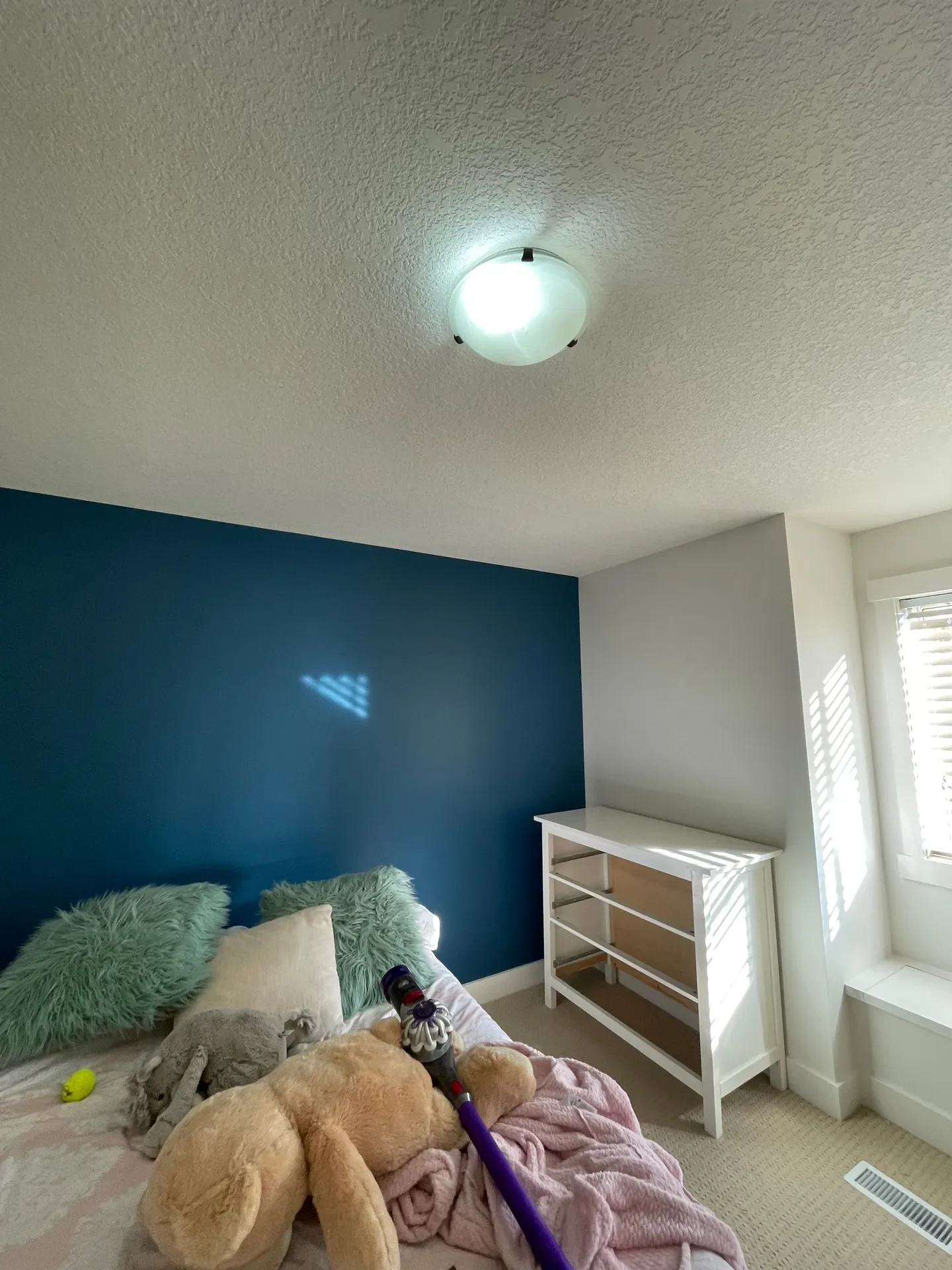 A bedroom with a blue accent wall, bed with pillows and plush toys, a vacuum cleaner, and natural light coming through the windows.