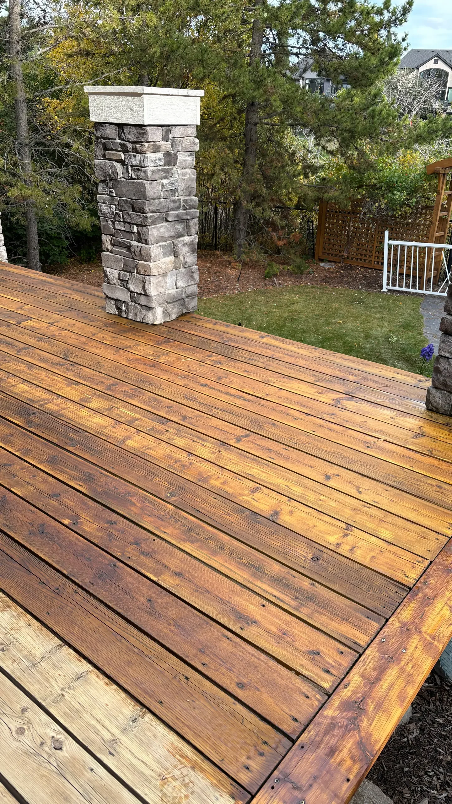 A wooden deck with a stone chimney, outdoor trees, and a backyard with grass, bushes, and part of a house with an outdoor staircase.