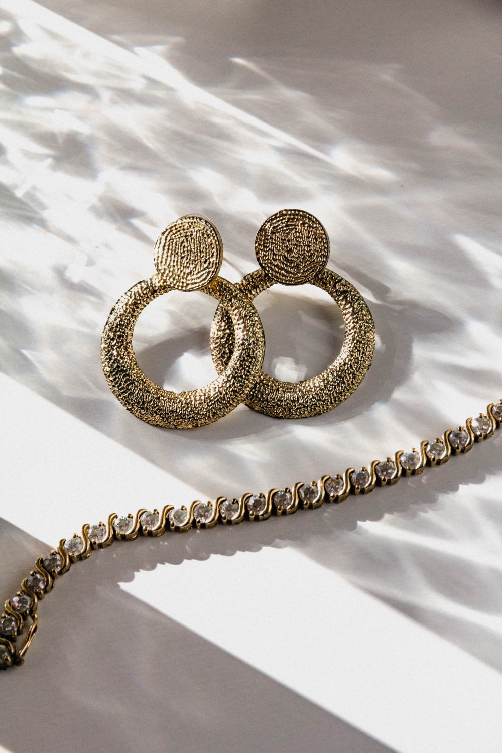 Gold textured hoop earrings with round top studs placed on a white surface with shadows cast by light, along with a gold necklace with clear gemstones.