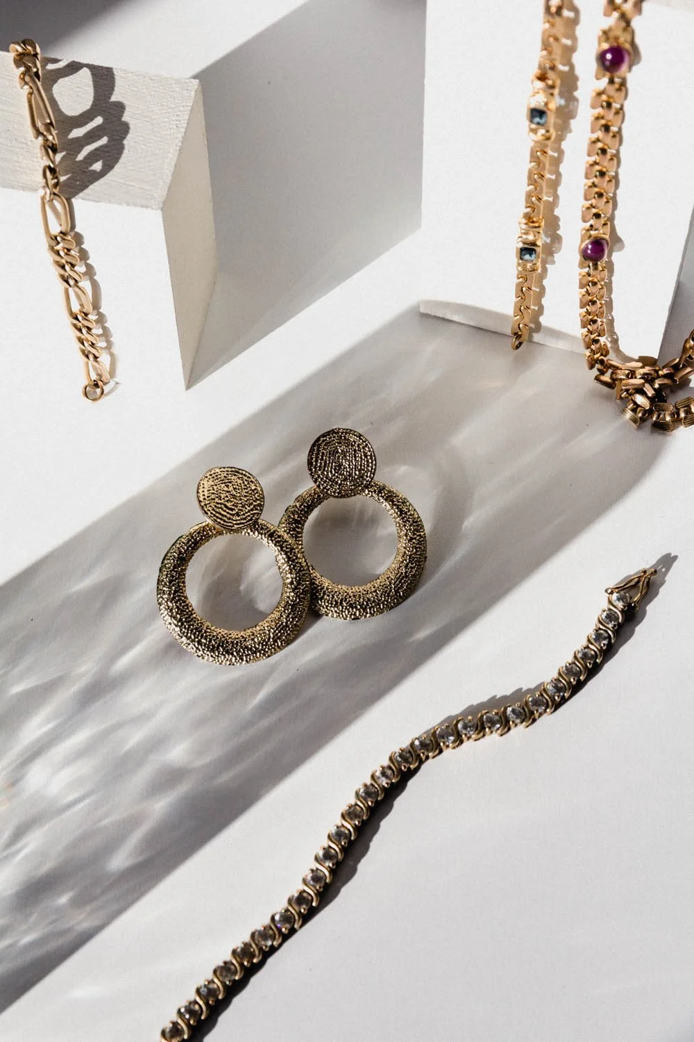 Various gold jewelry pieces including a chain, earrings, and necklaces displayed on a white surface with shadows.