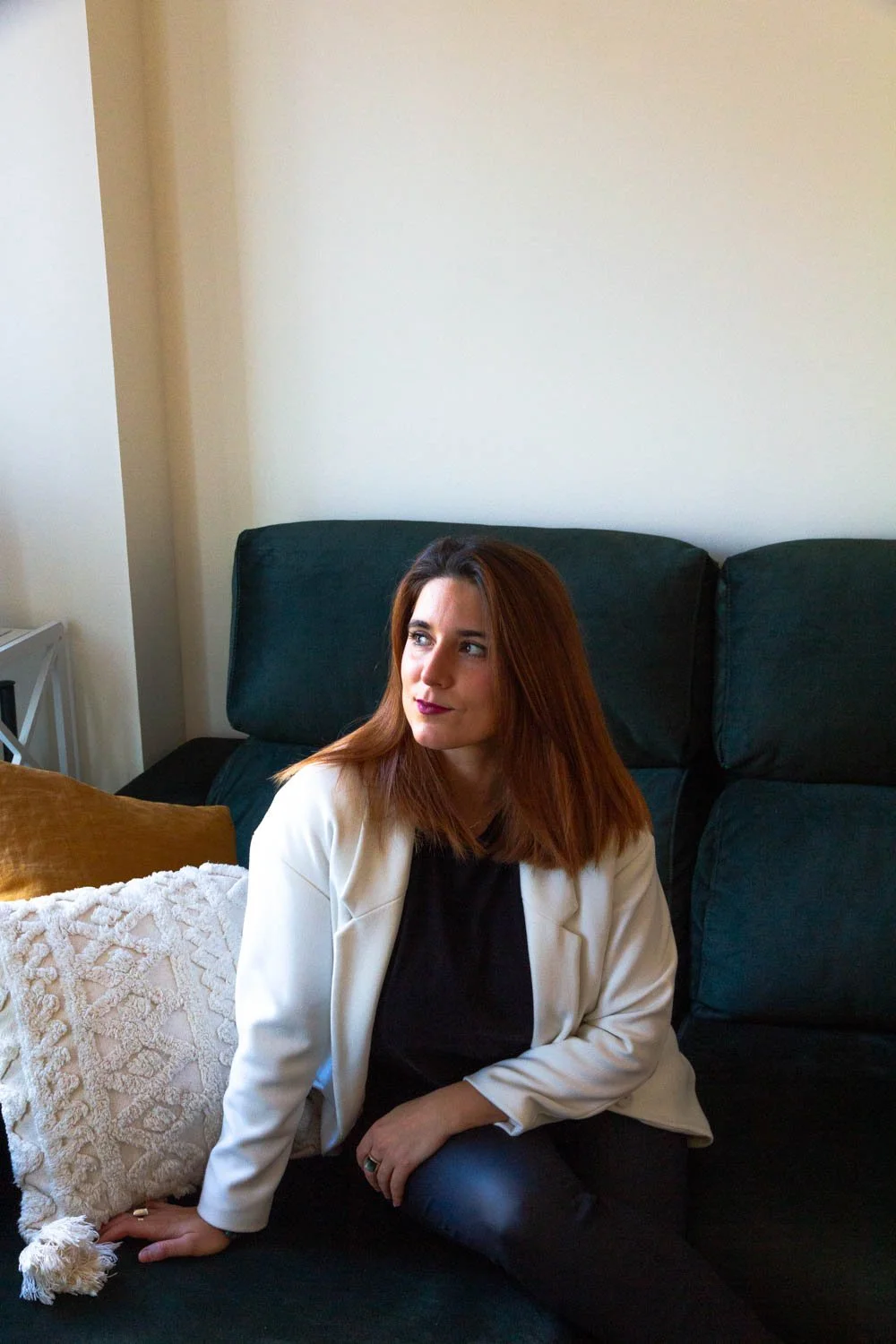 A woman with reddish-brown hair sitting on a dark green sofa, wearing a black top and a white blazer, looking to her right in a room with a white wall background.