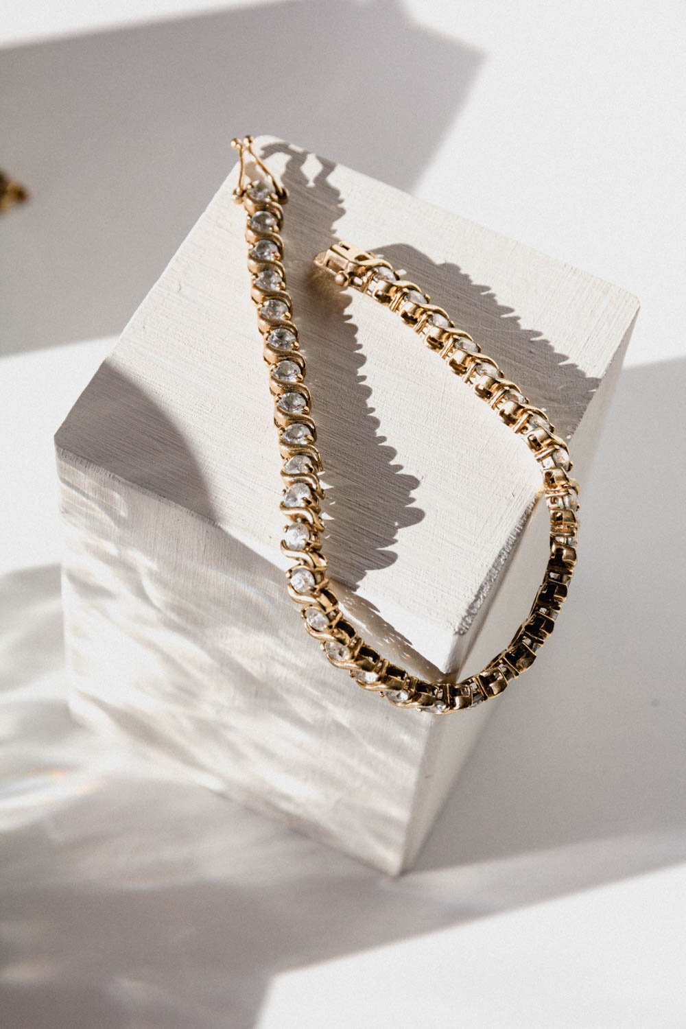 Gold bracelet with clear gemstones resting on a white textured block with shadows cast on a white background.