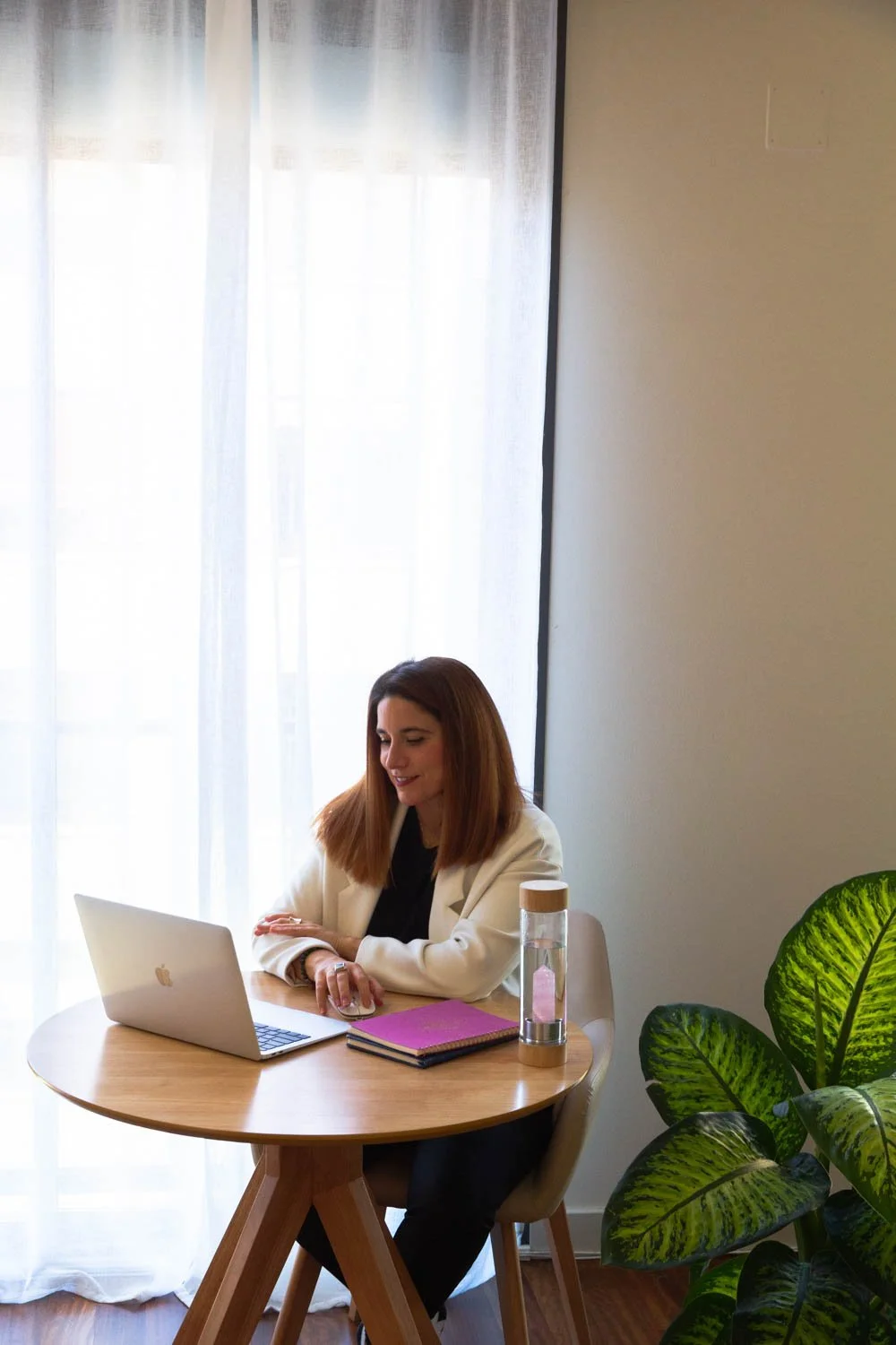 Woman with shoulder-length brown hair sitting at a wooden table with a silver laptop, purple notebook, and a water bottle, smiling while looking at her laptop in a room with sheer curtains, a green plant, and beige walls.