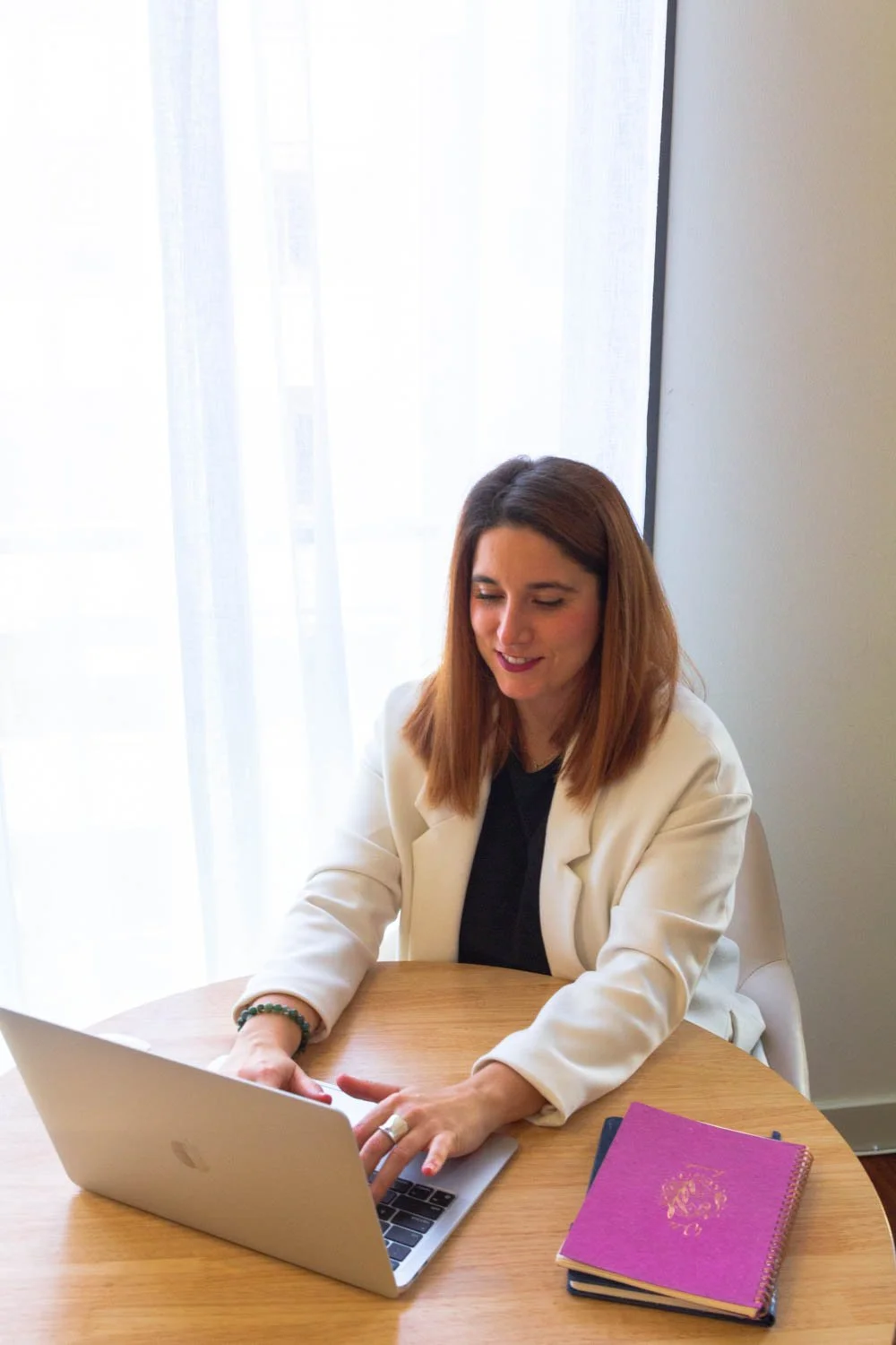 A woman with shoulder-length brown hair sits at a round wooden table, working on a silver laptop. She is wearing a white blazer and a dark top, smiling slightly. There is a purple notebook with a gold design beside her. Behind her, sheer white curtains cover a large window, letting in soft natural light.