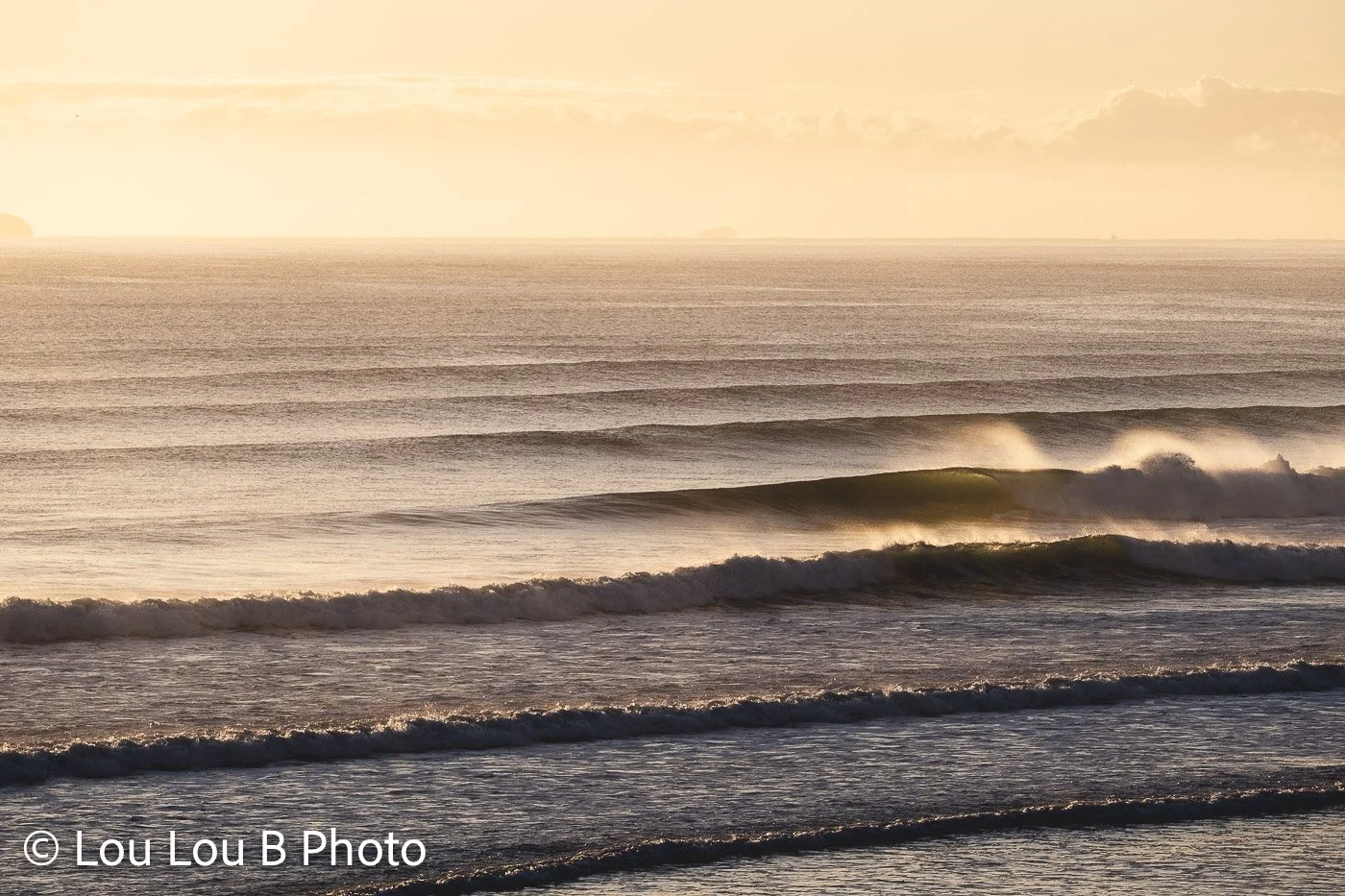 Lines of ocean swell glowing in warm evening light with spray blowing from the breaking waves.