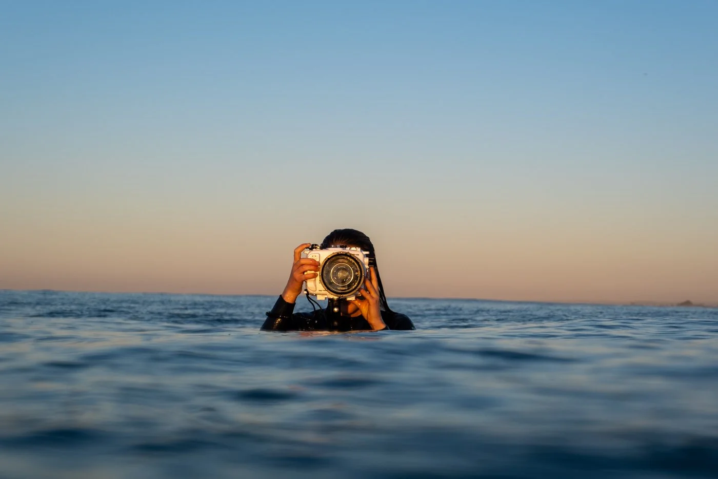 Surf photographer using a mirrorless camera in a surf photography water housing at ocean surface during sunset