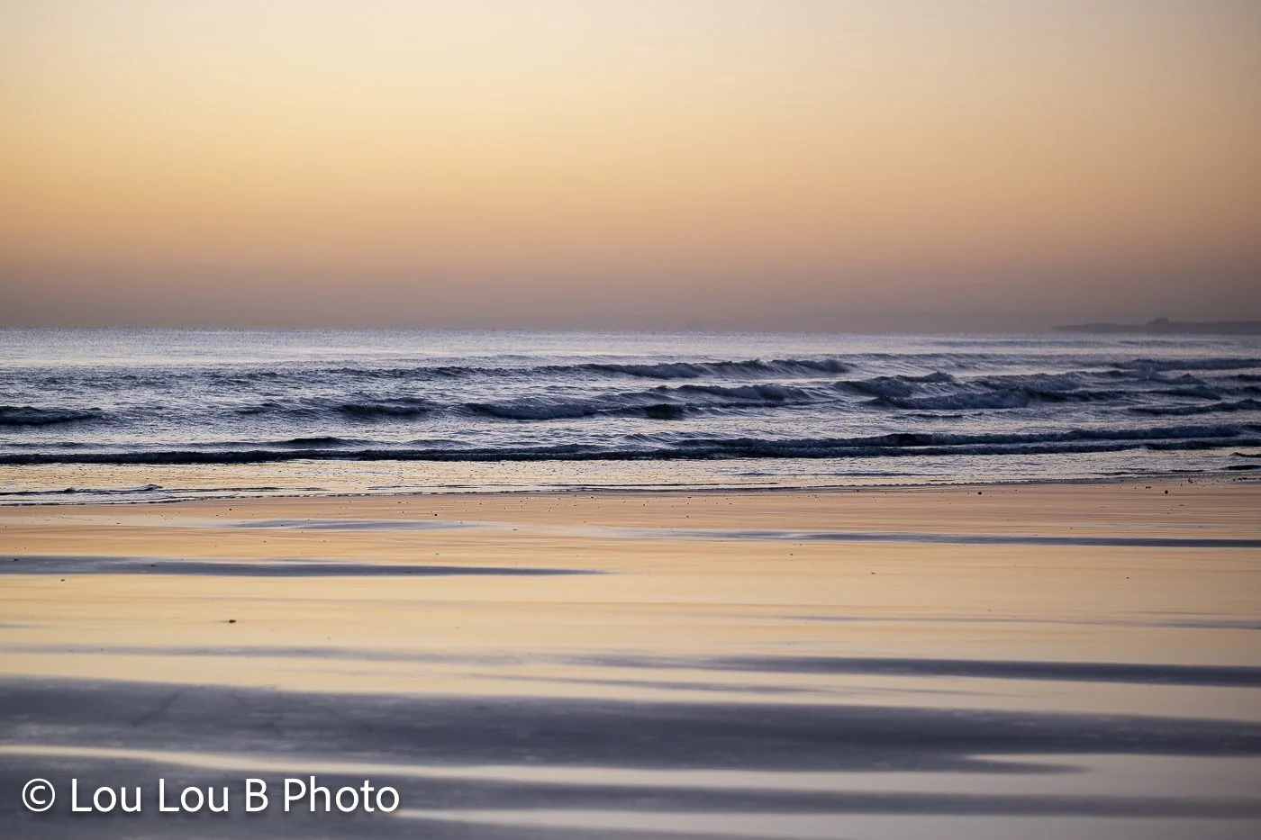Quiet beach and ocean beneath a peach and champagne-coloured sunset sky.