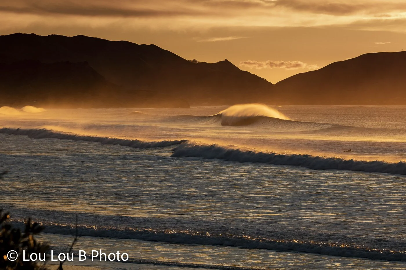 Ocean swell breaking in golden evening light beneath dark silhouetted coastal hills.