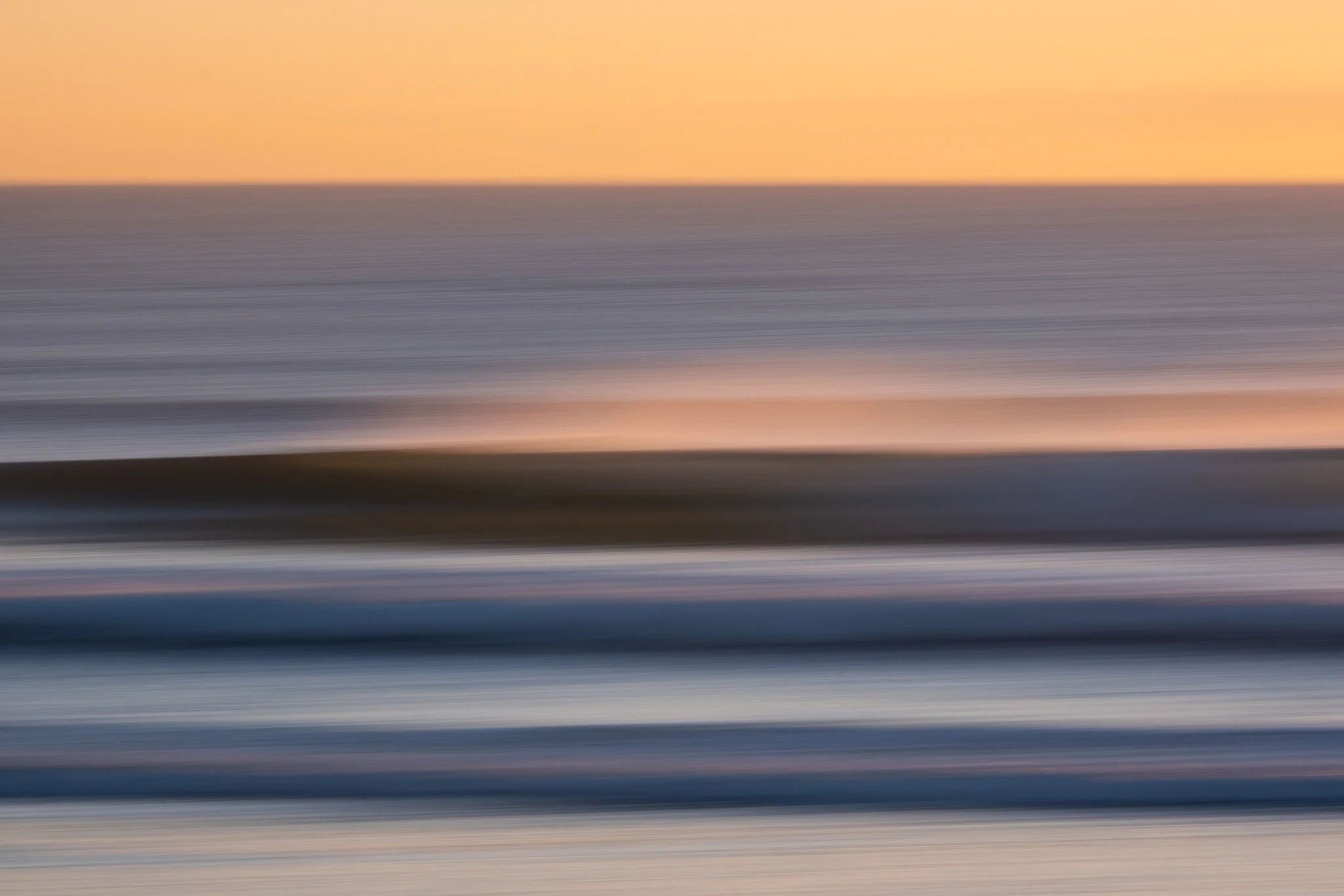 Stacked waves at sunrise in Mount Maunganui, New Zealand. The photo is taken using slow shutter and panning technique, creating a sense of motion and emphasizing the warm tones of sunrise and blues of the ocean. A beautiful surf lifestyle photo by Lo