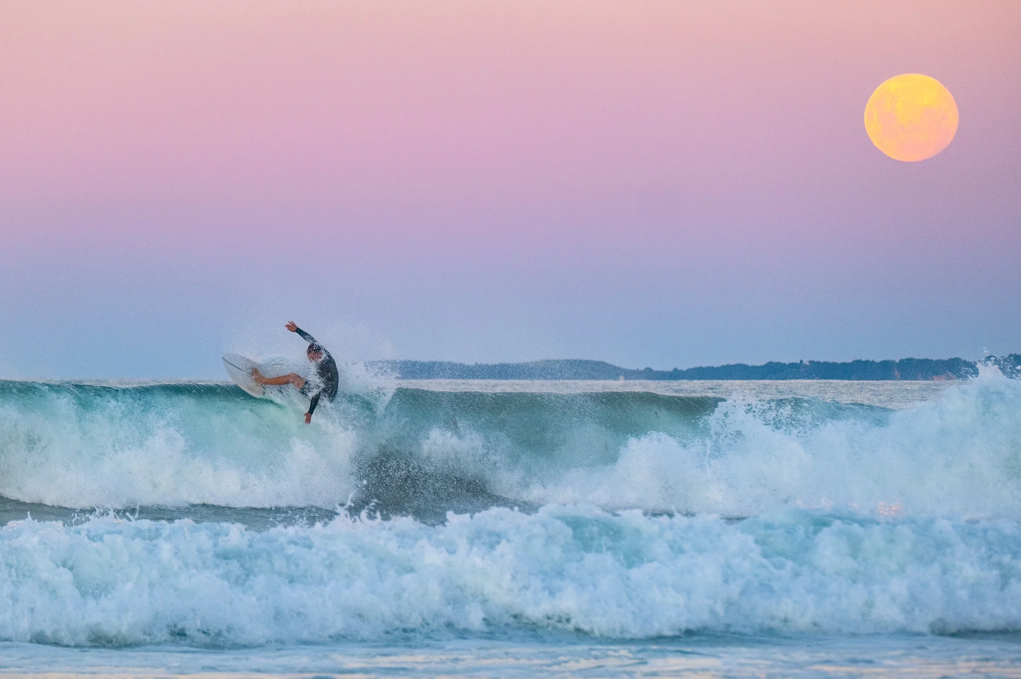A surfer rides the crest of a wave just before it breaks, with pastel hues of pink and orange filling the sky at sunset. A giant moon rises in the background, casting a warm glow on the water below.