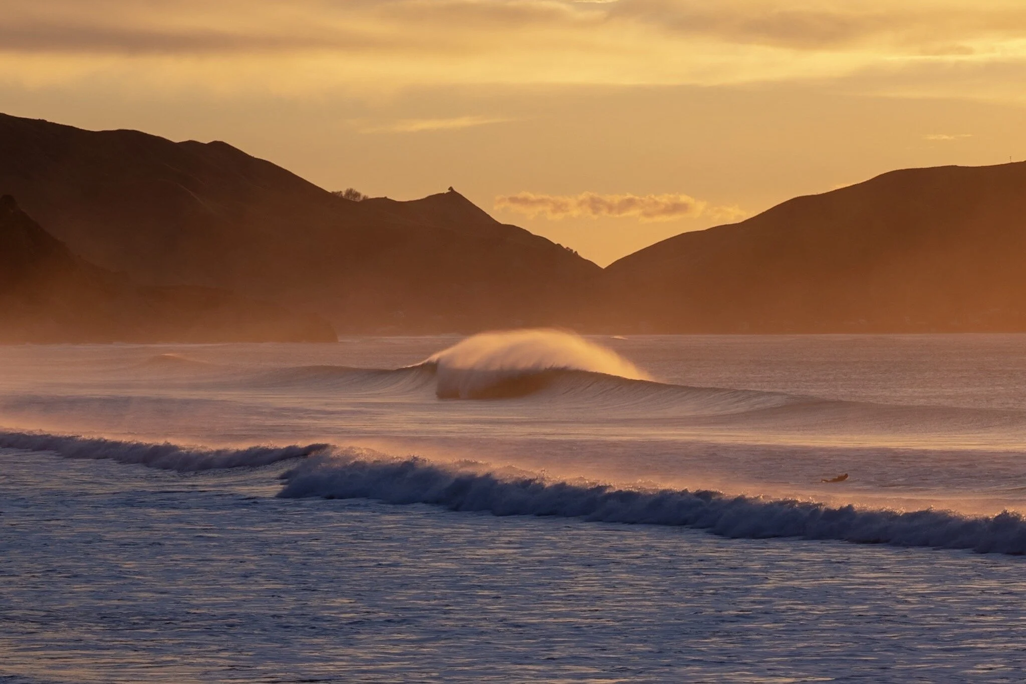 Sunrise at Wainui Beach, Gisborne. A perfect wave breaks and barrels in the foreground, with warm orange light bathing the scene. Cliffs silhouette in the background, adding to the landscape composition. Lou Lou B Photo captures the power and beauty 
