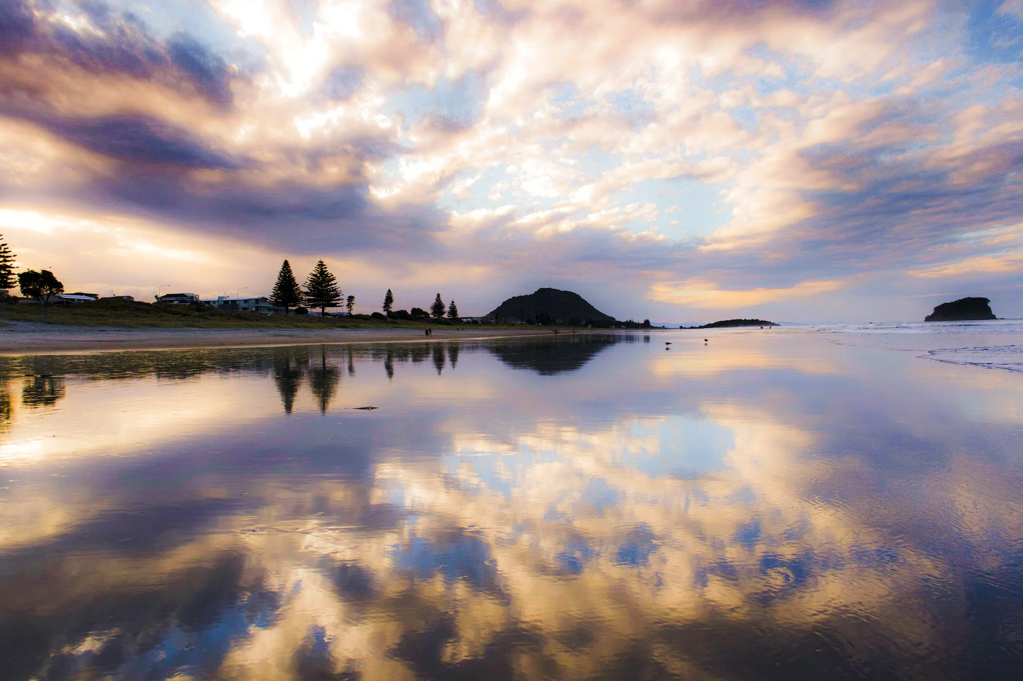 Mount Maunganui at low tide, with the whole beach scene reflected in the wet sand. The sky is a mixture of blues and purples with clouds stretching across the frame. Wide angle lens used to capture the expansive view. Beach landscape photo by Lou Lou