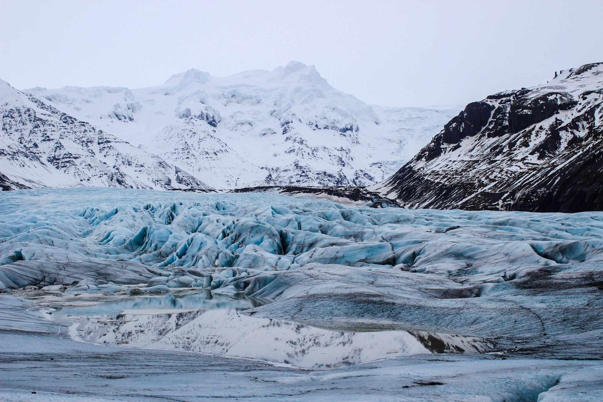 A stunning image of a glacier in Iceland. The glacier is a mix of shades of white and blue, with turquoise pops of color adding to the visual interest. The surrounding mountains provide a majestic backdrop, with their peaks shrouded in mist. The phot