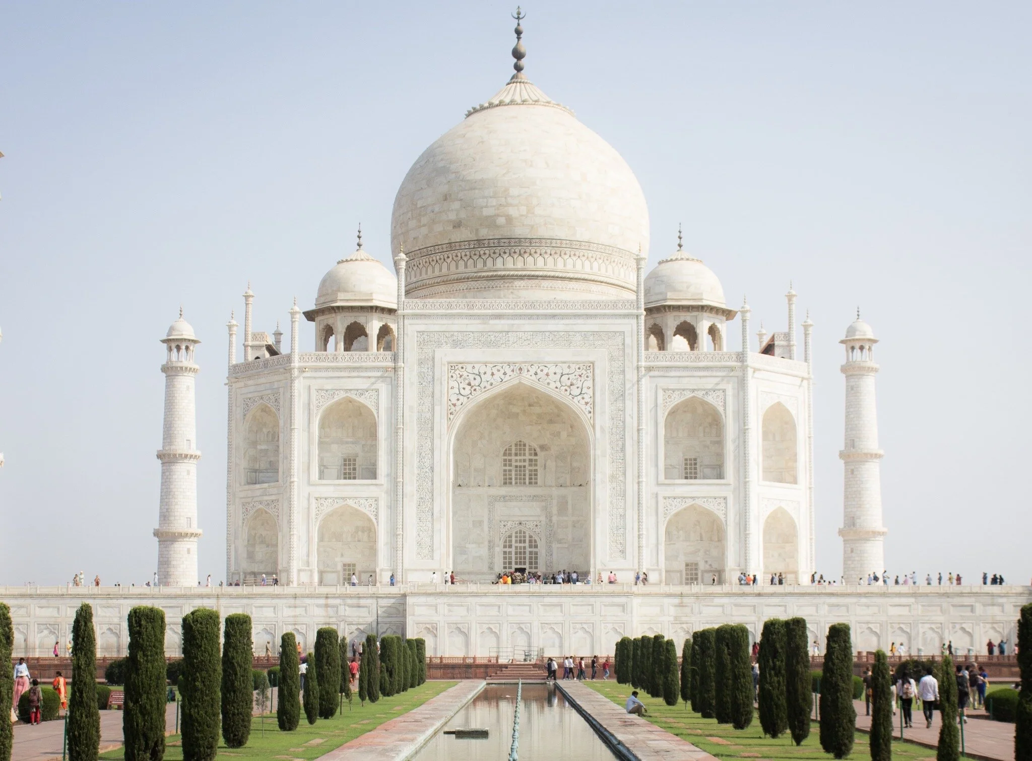 A stunning view of the Taj Mahal in Agra, India. The white marble mausoleum is reflected in a calm body of water in front of it, with the clear blue sky in the background. The intricate details of the architecture are visible, including the ornate do