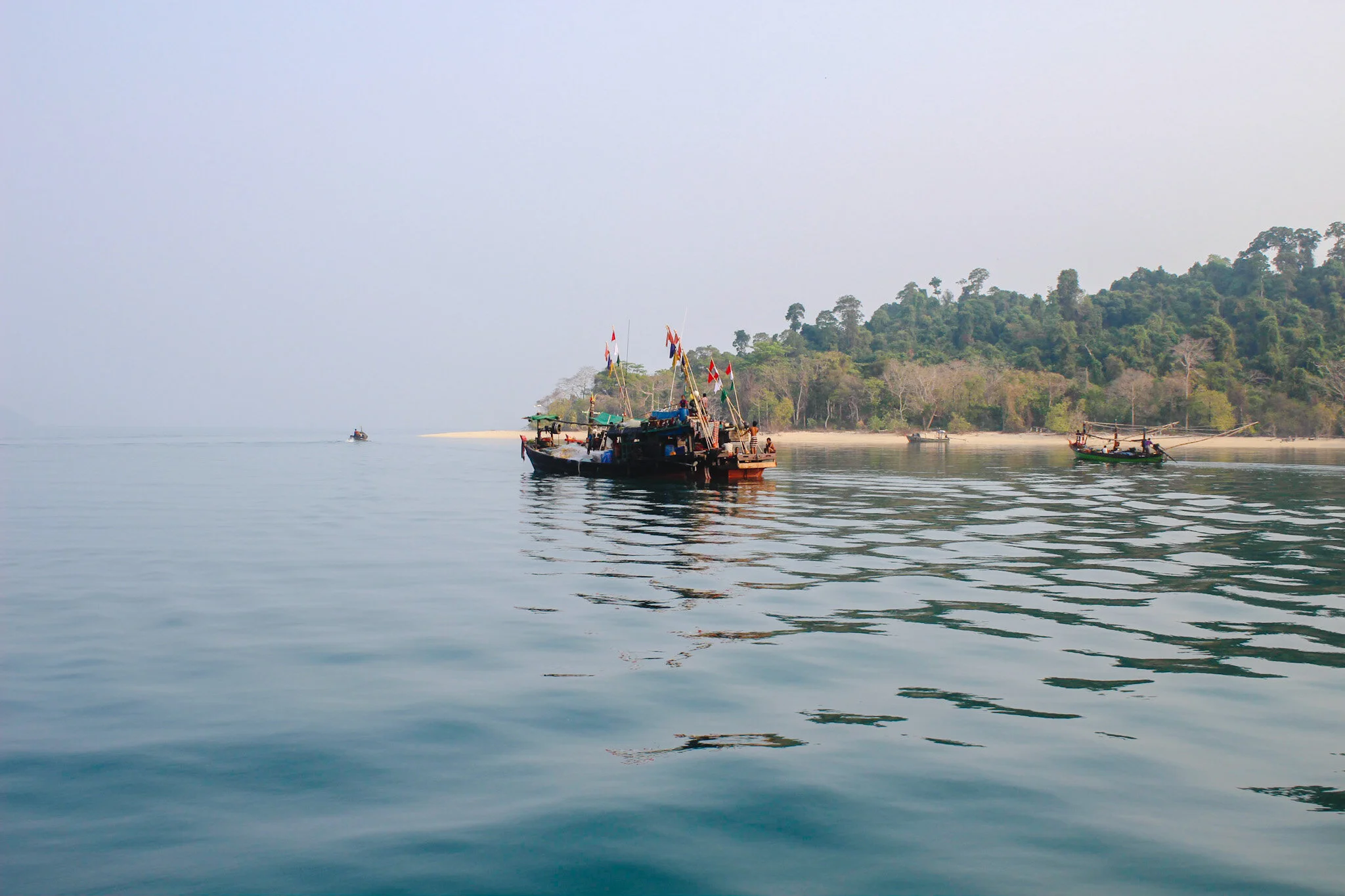 A picturesque view of a tropical island in Myanmar. In the foreground, a traditional boat and local people can be seen, while the background shows a lush green island with white sandy beaches and clear turquoise waters. The photo was taken from the s