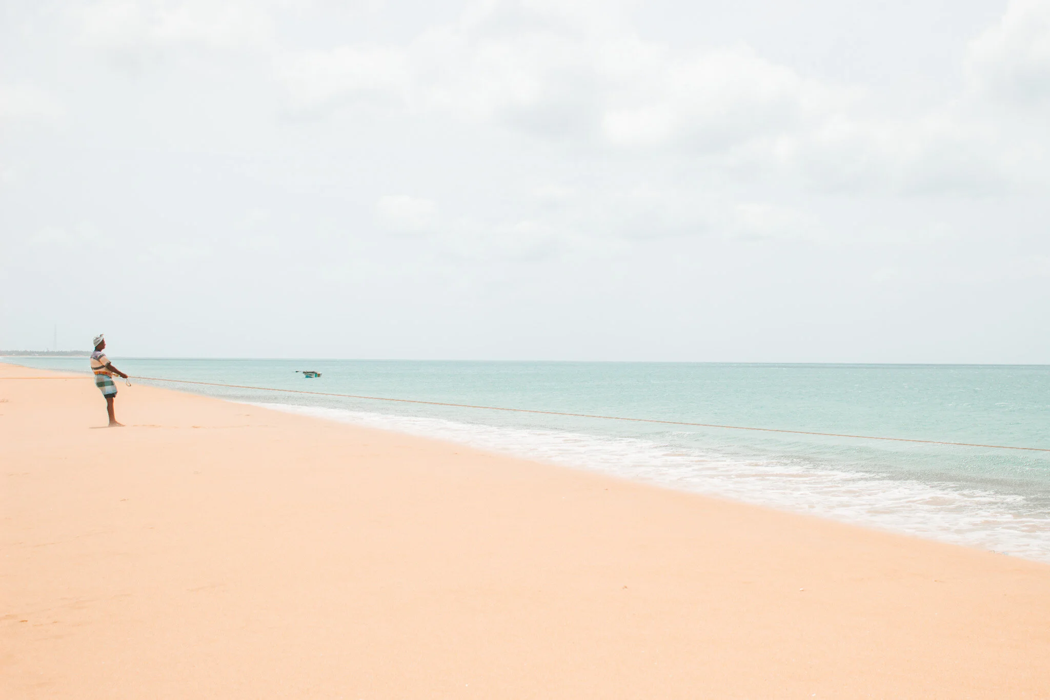 A serene and tranquil image of a deserted beach in Sri Lanka. The photo captures a lone fisherman standing at the edge of the water, pulling in his fishing net from the land. The clear turquoise sea stretches out in the background, creating a serene 