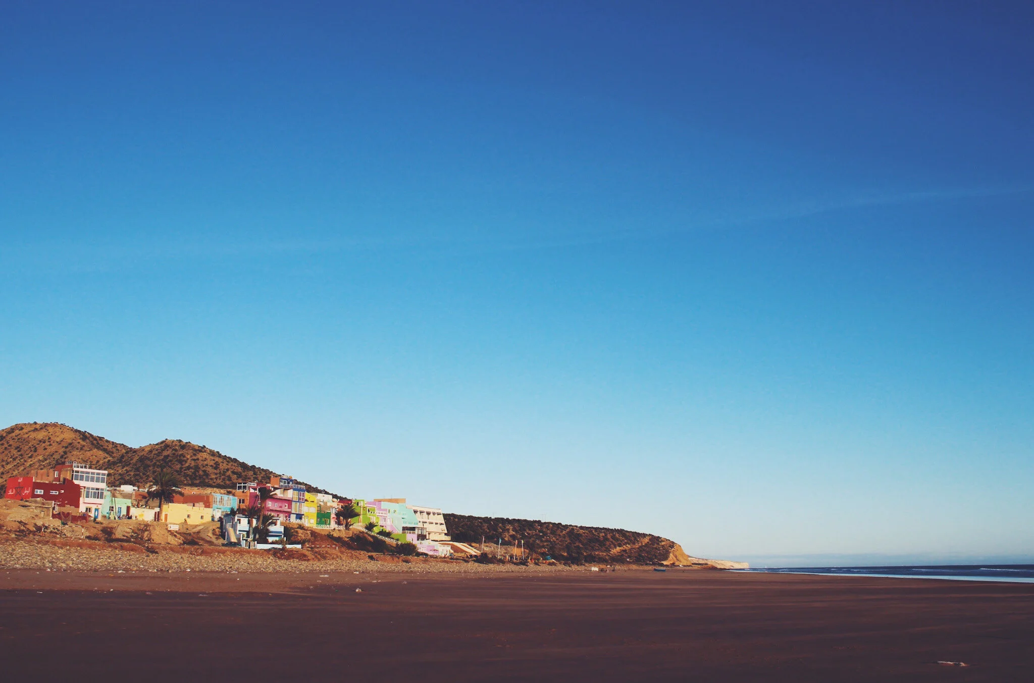 A beautiful view of a Moroccan beach. The photo shows a long stretch of golden sand leading up to the calm waters of the sea. In the distance, vibrant houses and buildings can be seen, adding a splash of color to the scene. The clear blue sky overhea