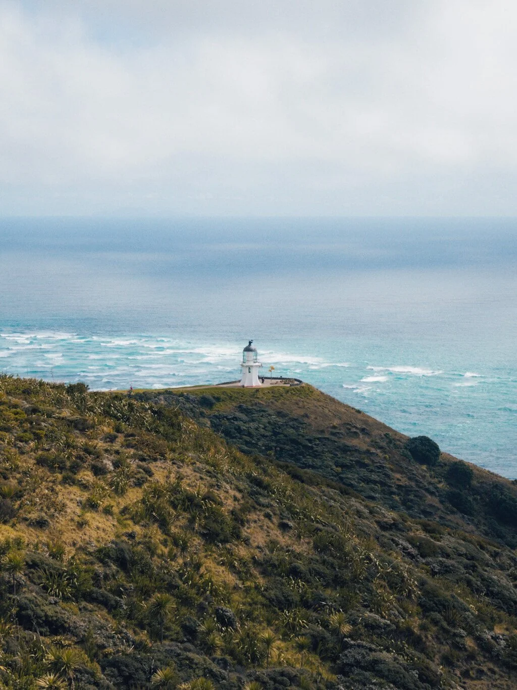 A stunning view of Cape Reinga Lighthouse, situated in the lush green landscape of New Zealand. The lighthouse stands tall in the foreground, overlooking the vast turquoise ocean where the Tasman Sea and Pacific Ocean meet. The powerful collision of 