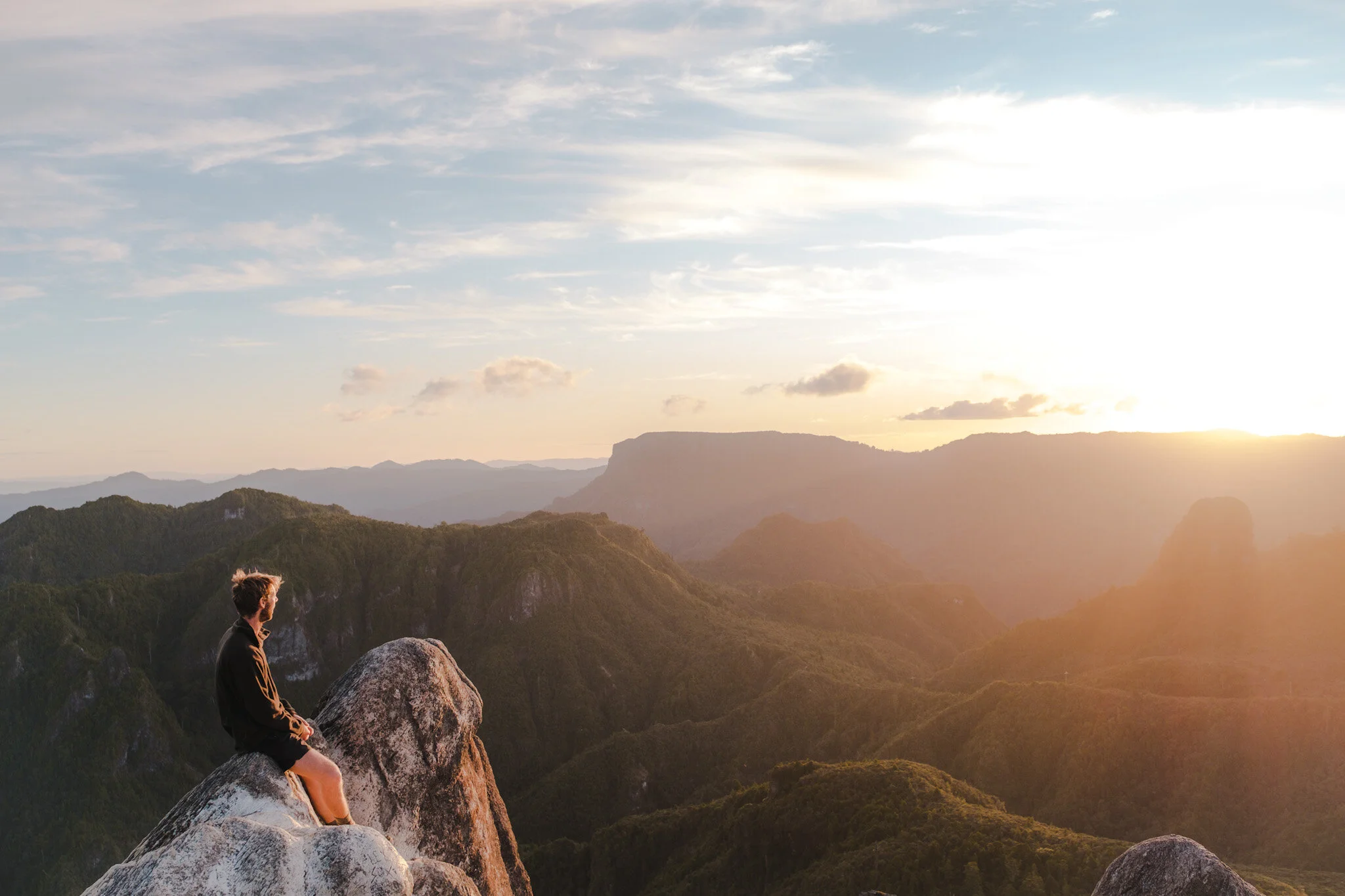 Experience the breathtaking beauty of New Zealand's Coromandel with this stunning photograph taken from the summit of the Pinnacles hike. The image captures the vibrant hues of the sunset as a man sits atop a rock, taking in the panoramic view. The r