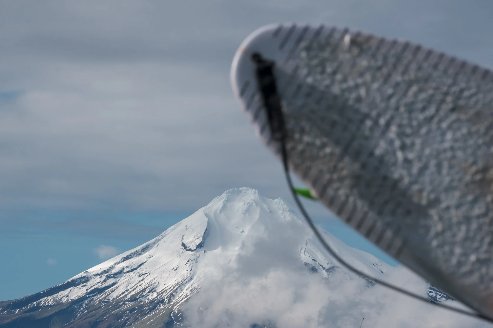 Mount Taranaki, snow-capped in the background, with the tail of a surfboard in the foreground, a perfect surf lifestyle photo capturing the essence of this region. Photo by Lou Lou B Photo.