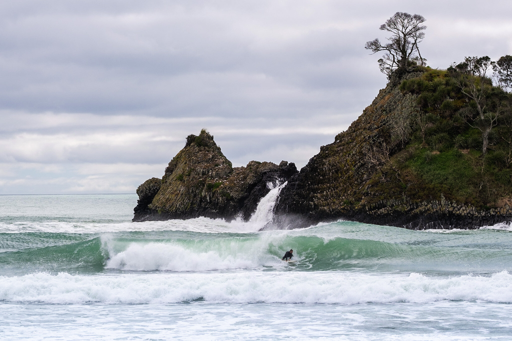Surfer rides barrelling wave on side of the peak, breaking in front of a rocky outcrop at Whangapoua Beach in the Coromandel, captured from the land by Lou Lou B Photo. The blue-green water crashes into white foam as the surfers carve their way throu