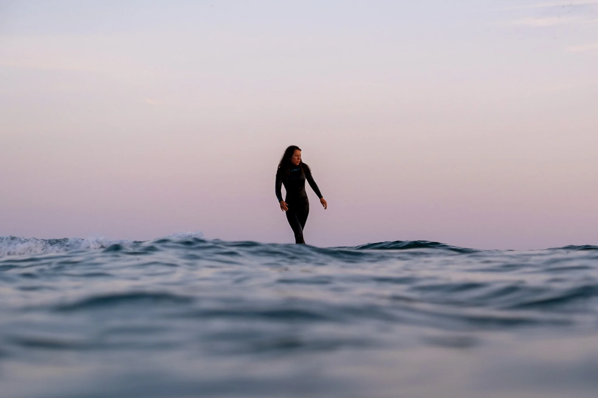 A woman surfs on a longboard in a black wetsuit during dawn, with a pastel purple sky and blue ocean behind her. Surf lifestyle photo by Lou Lou B Photo.